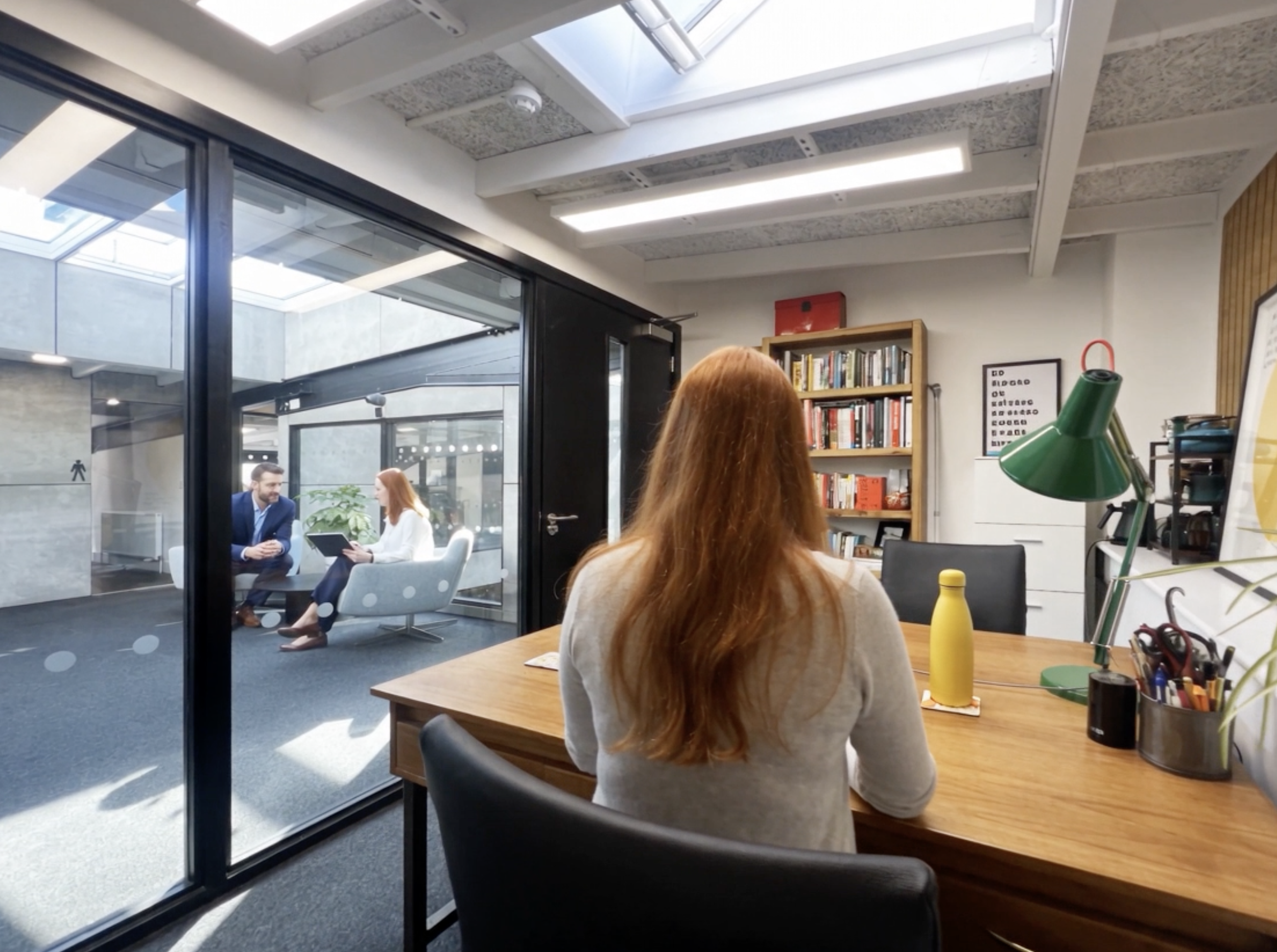 Inside an office with a woman with long red hair sitting at a wooden desk, facing away. In the background through glass doors, a man in a blue suit and a woman are seated in a bright lounge area, having a conversation. The office has a bookshelf filled with books, a framed word list on the wall, a green desk lamp, and various office supplies on the desk.