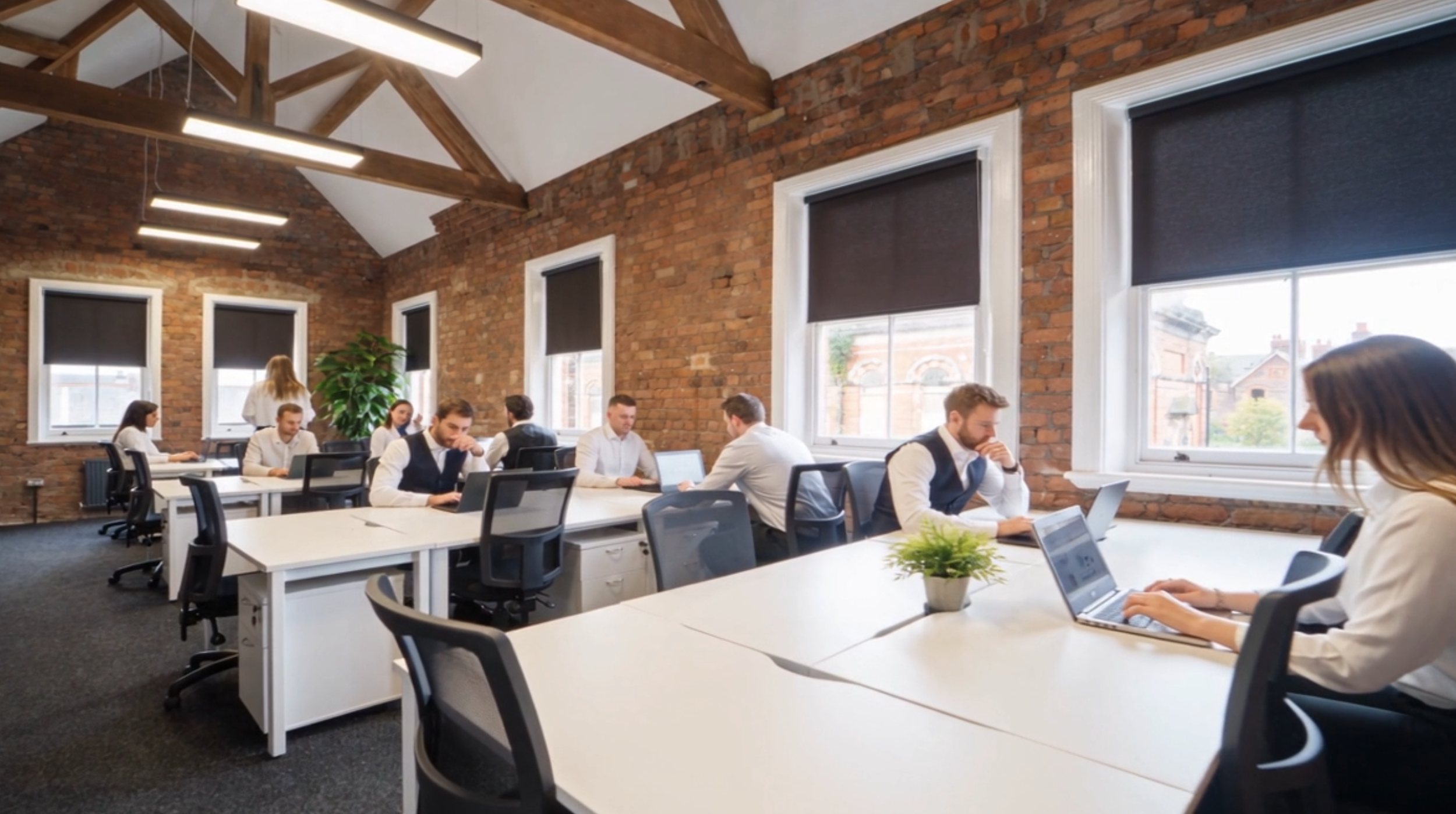 Office with exposed brick walls, large windows with dark blinds, wooden beams on the ceiling, and multiple people working on laptops at white desks.