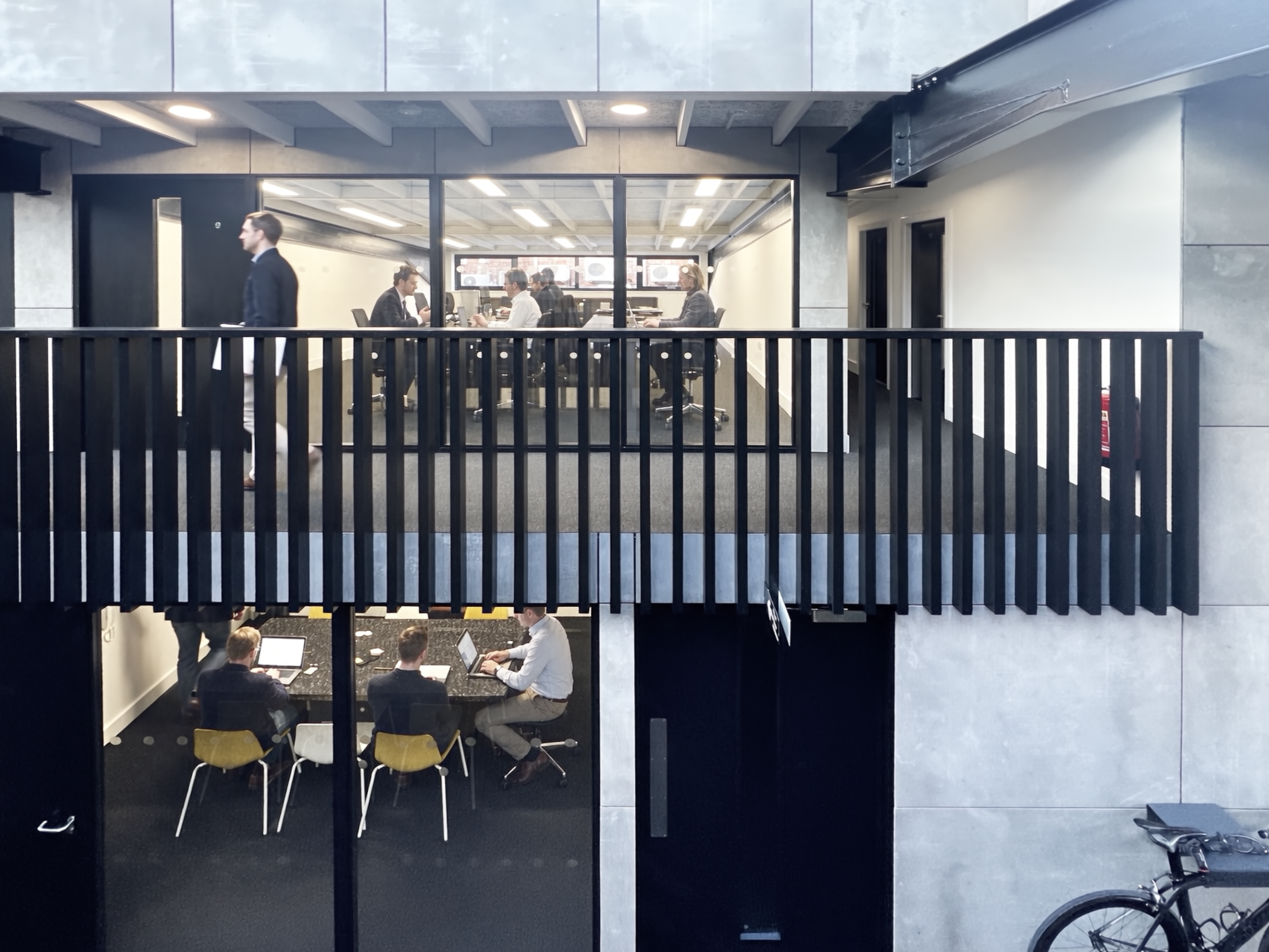 Office meeting room with people working on laptops and a man walking past. The room has large windows and modern decor.