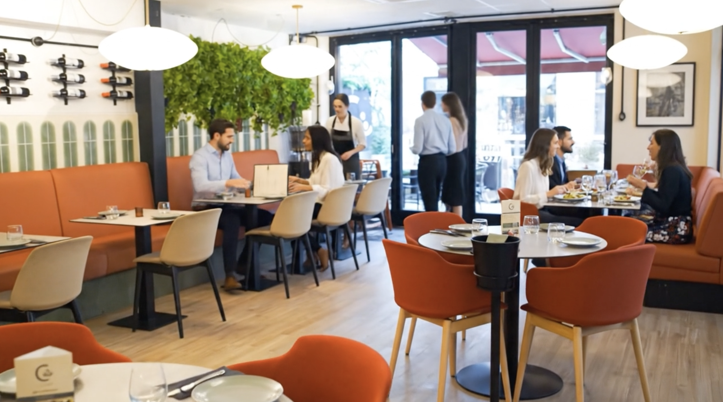 A cozy restaurant with several tables set for dining, including plates, glasses, and utensils. People are sitting and having conversations, while waitstaff serve customers. The interior features orange and beige seating, large windows, and decorative greenery.