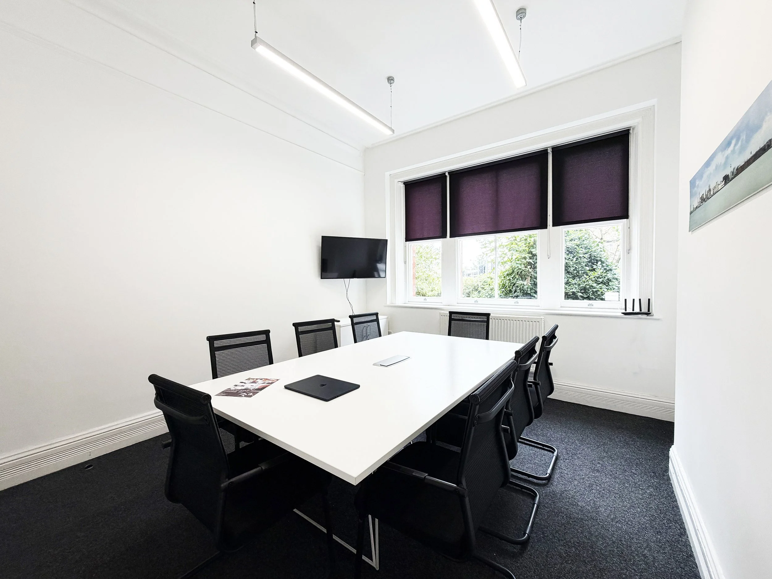 A modern conference room with a white rectangular table, black mesh office chairs, a wall-mounted television, and a large window with dark purple blinds, letting in natural light and greenery outside.