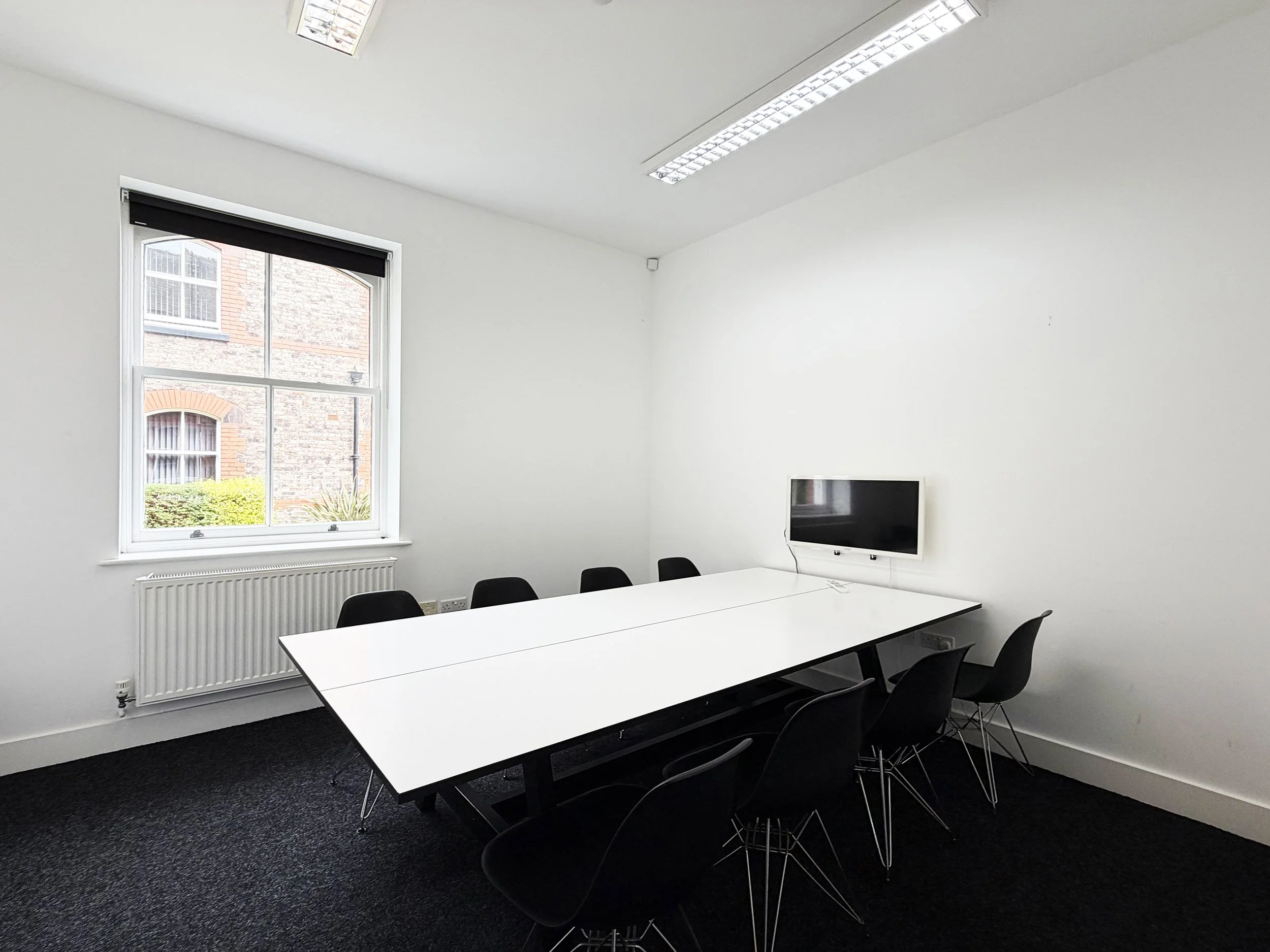 A modern conference room with white walls, a large window showing brick and stone buildings outside, a white radiator beneath the window, black chairs arranged around a white rectangular table, a flat-screen television on the wall, and fluorescent ceiling lights.