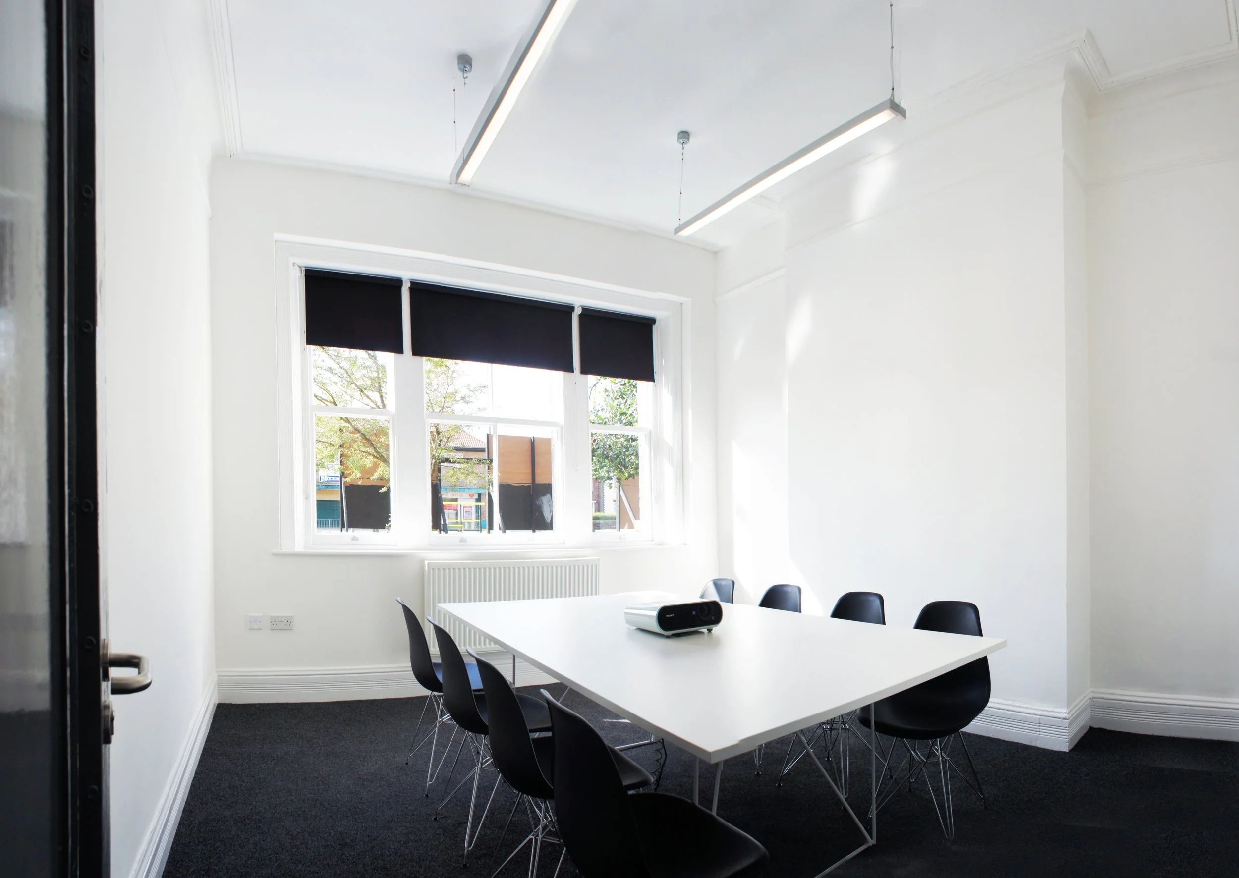 A modern meeting room with white walls, a white table, black chairs, large windows with black roller blinds, and a projector on the table.