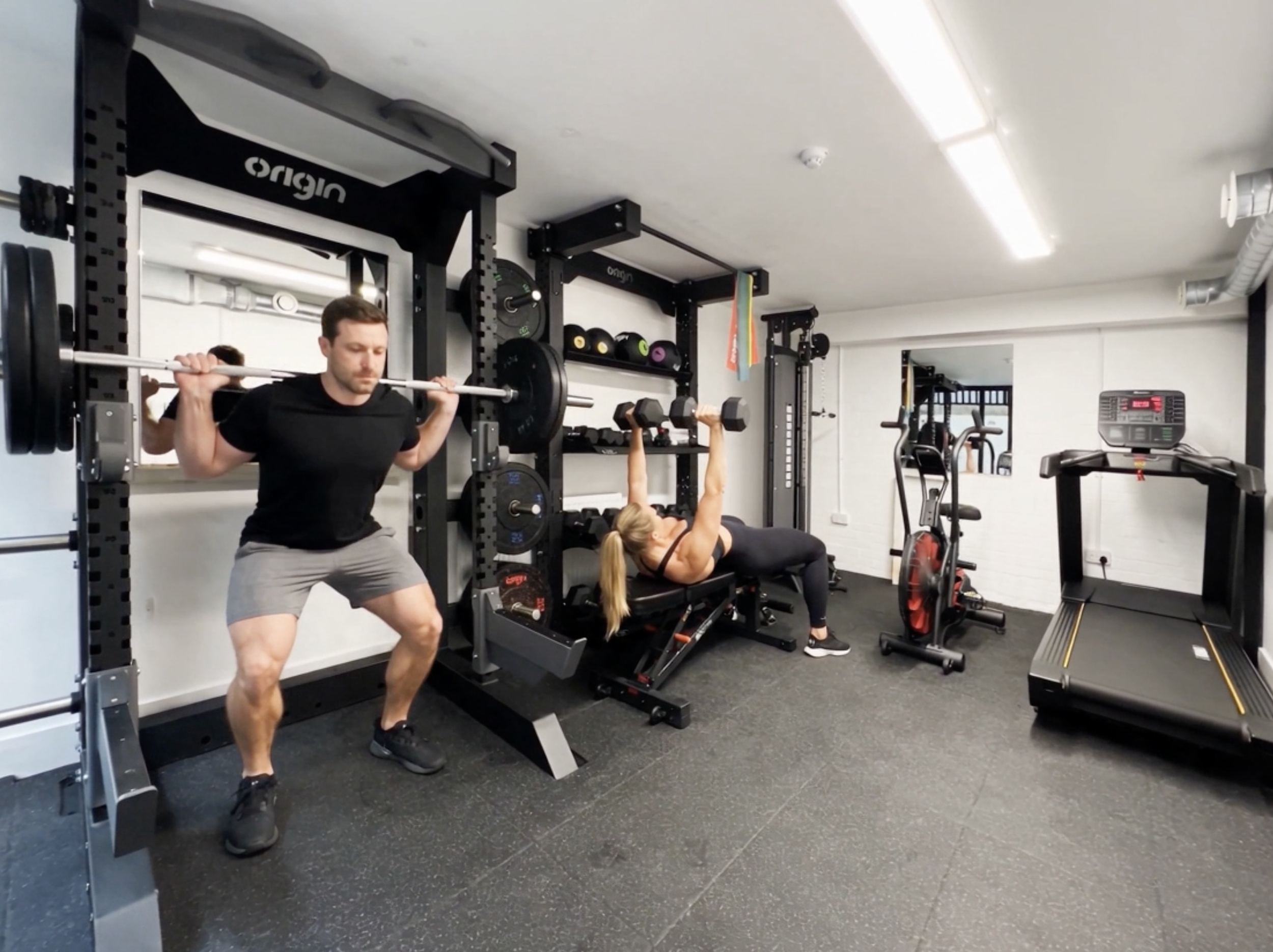 A man and a woman exercising in a gym, with the man lifting a barbell on his shoulders and the woman performing a sit-up or crunch on a workout bench.