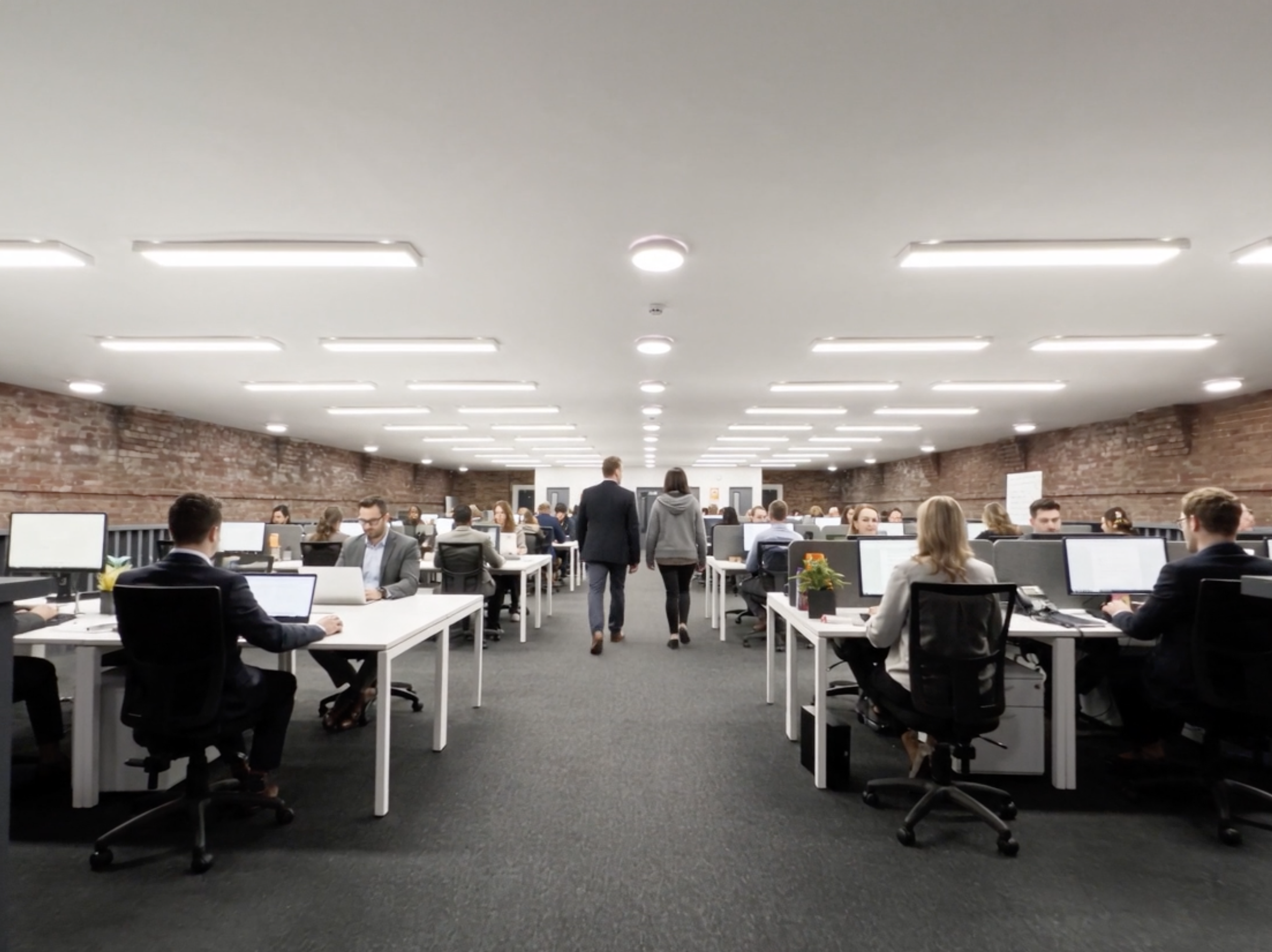 Large open office space with rows of desks, many people working on computers, exposed brick walls, and a drop ceiling with fluorescent lights.