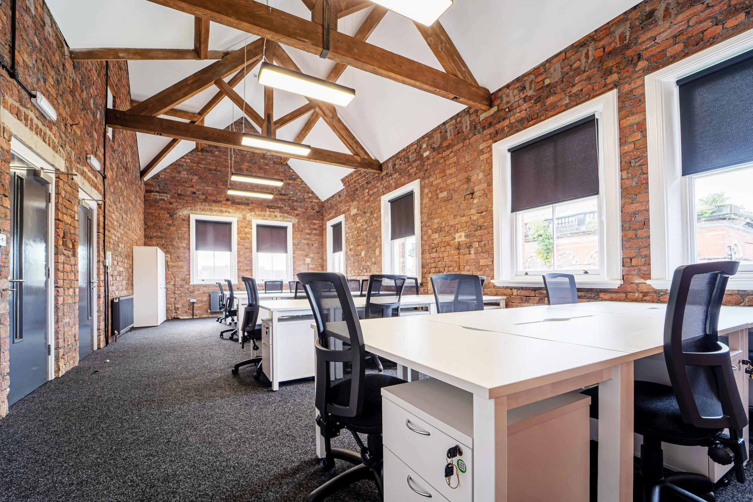 An office space with exposed red brick walls, large windows with black blinds, and a ceiling with wooden beams. There are white desks and black office chairs arranged in rows, with a gray carpeted floor and modern LED light fixtures hanging from the ceiling.