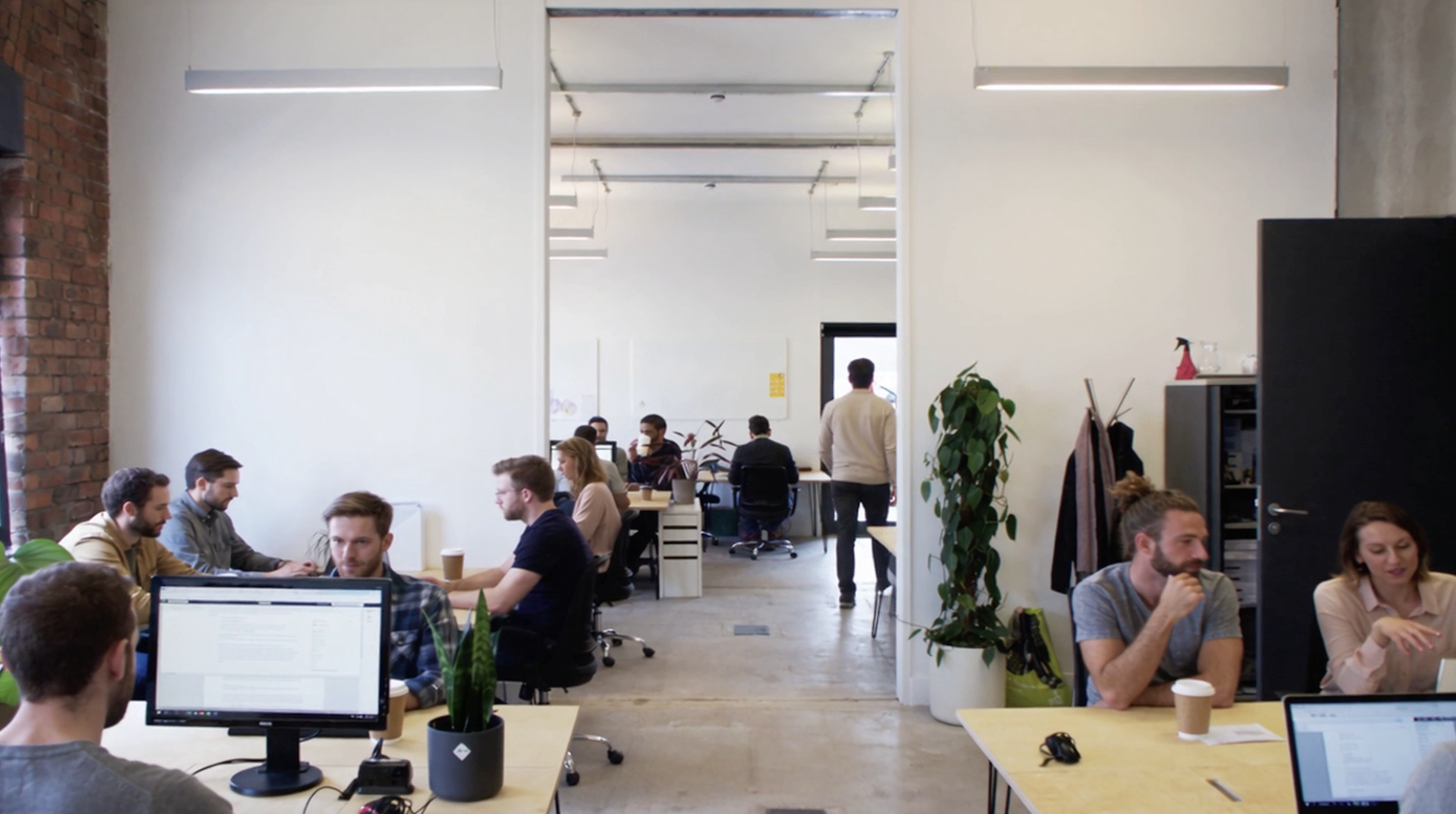Open-plan office with multiple people working at desks, some using computers, in a modern space with white walls, a brick wall on the left, and hanging light fixtures. A large white wall section and a doorway in the back reveal more seating and people working.