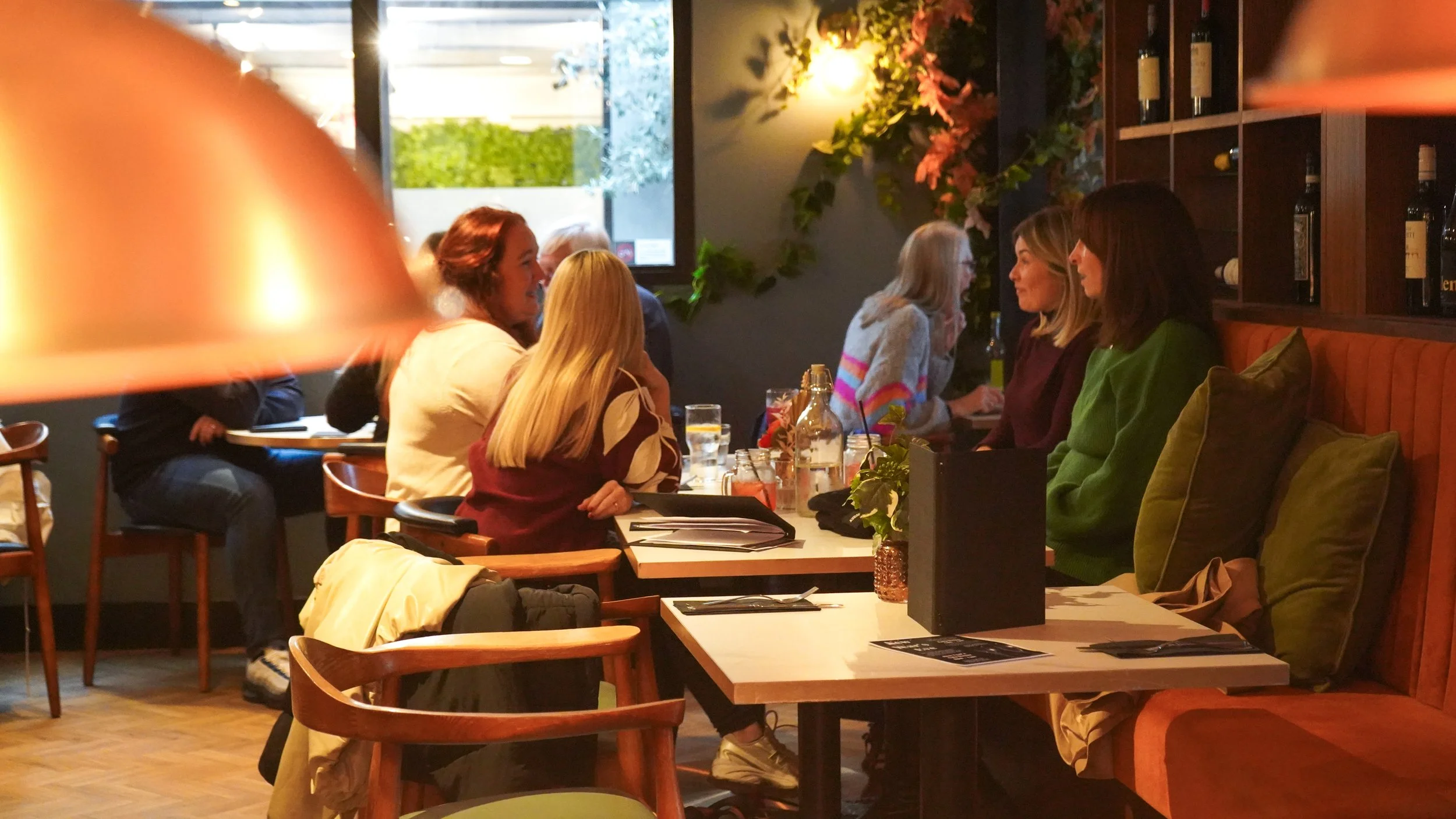 People sitting and talking at tables inside a cozy restaurant decorated with flowers and warm lighting.