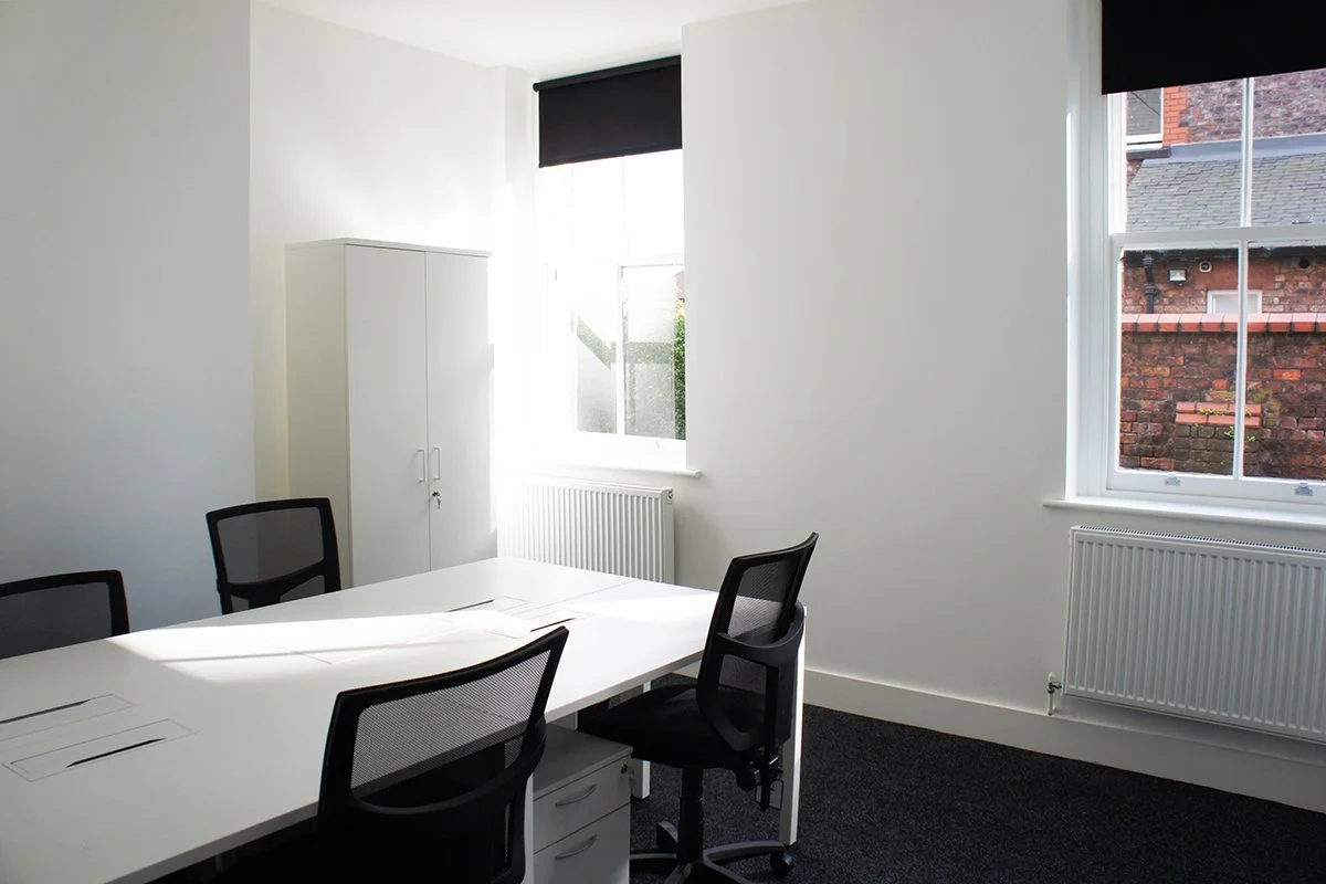 Empty modern office room with white walls, black window blinds, a white table with black mesh chairs, a white cabinet, and two radiators under the windows.