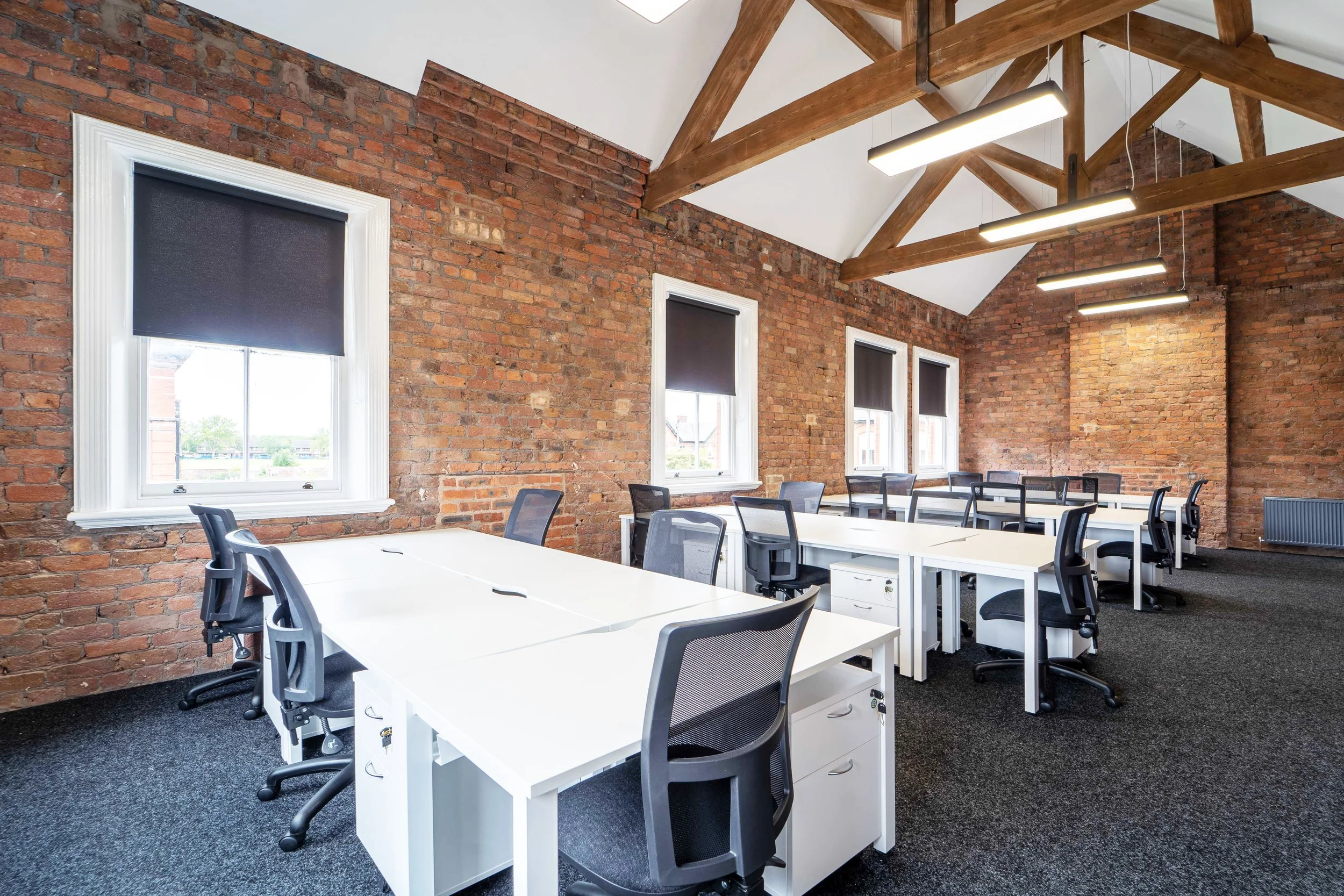 Modern office conference room with white desks, black chairs, exposed brick walls, large windows with dark blinds, and wooden ceiling beams.