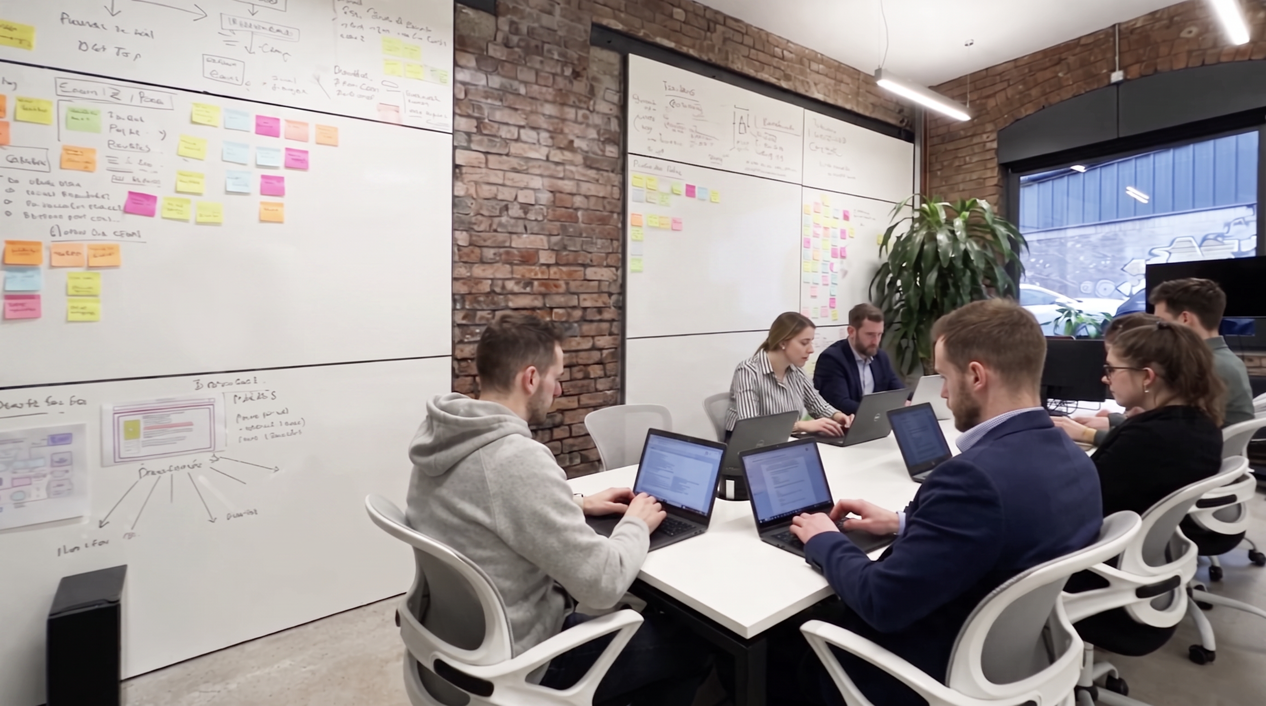 A group of people in a meeting room working on laptops, with whiteboards covered in notes and sticky notes on the wall, brick walls, and large window showing an urban exterior.