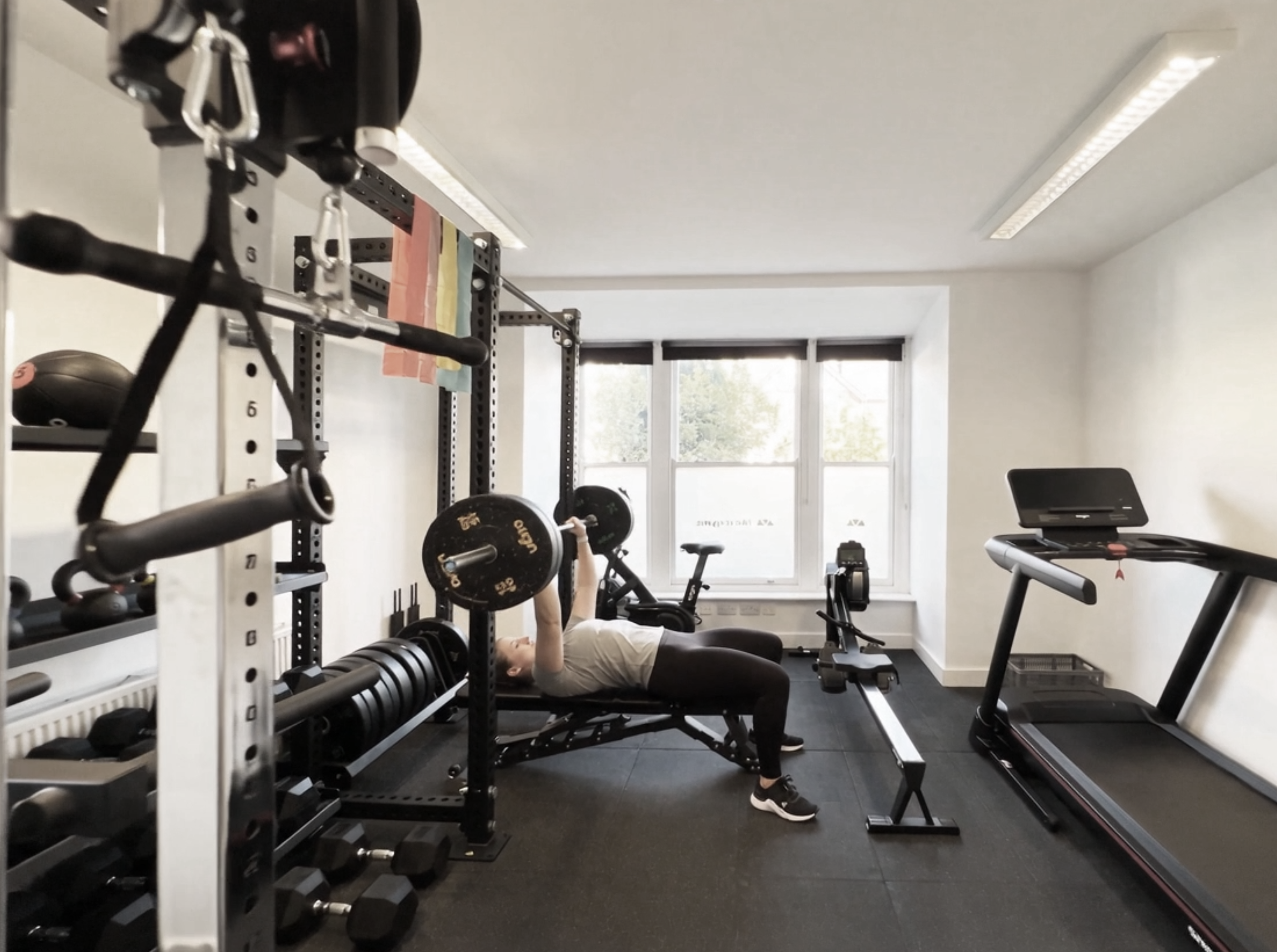 A person lying on a workout bench lifting a barbell in a gym, surrounded by various weights, gym equipment, and a bright room with large windows.