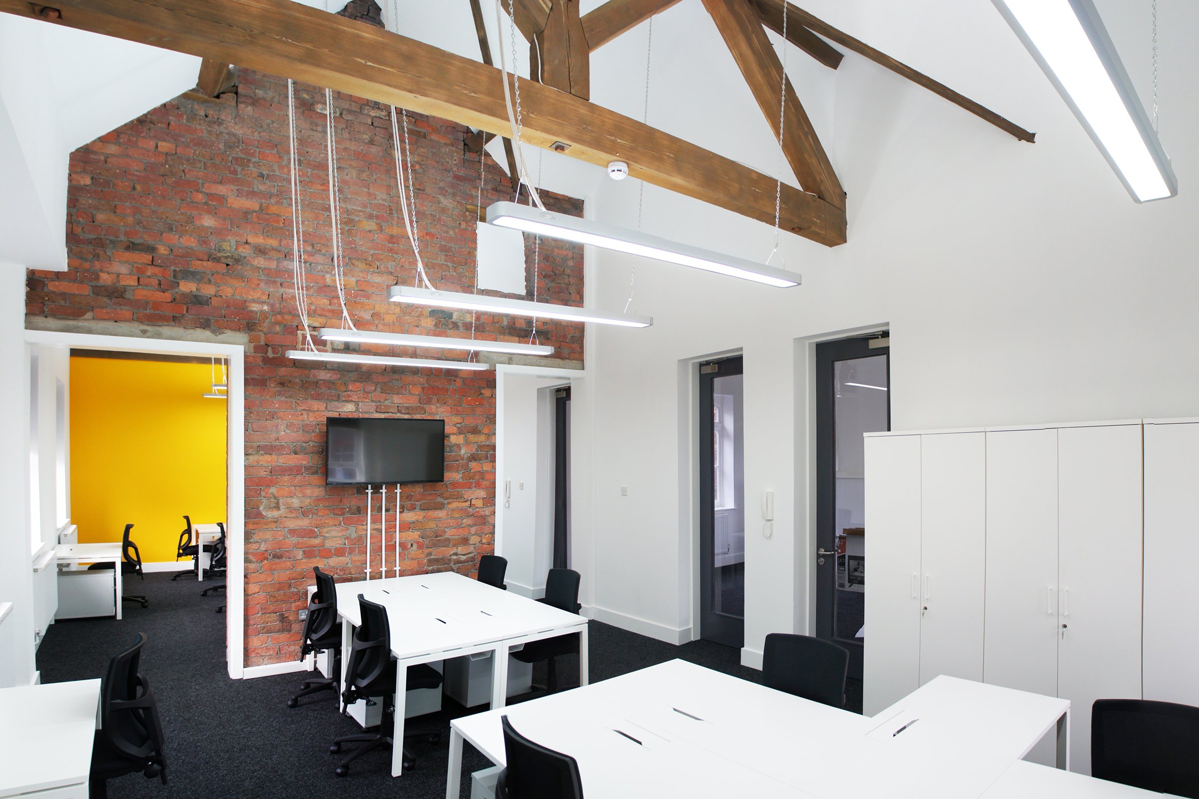 Modern office space with white tables, black chairs, a brick accent wall, a flat-screen TV, and multiple bright overhead lights hanging from a wooden ceiling beam.