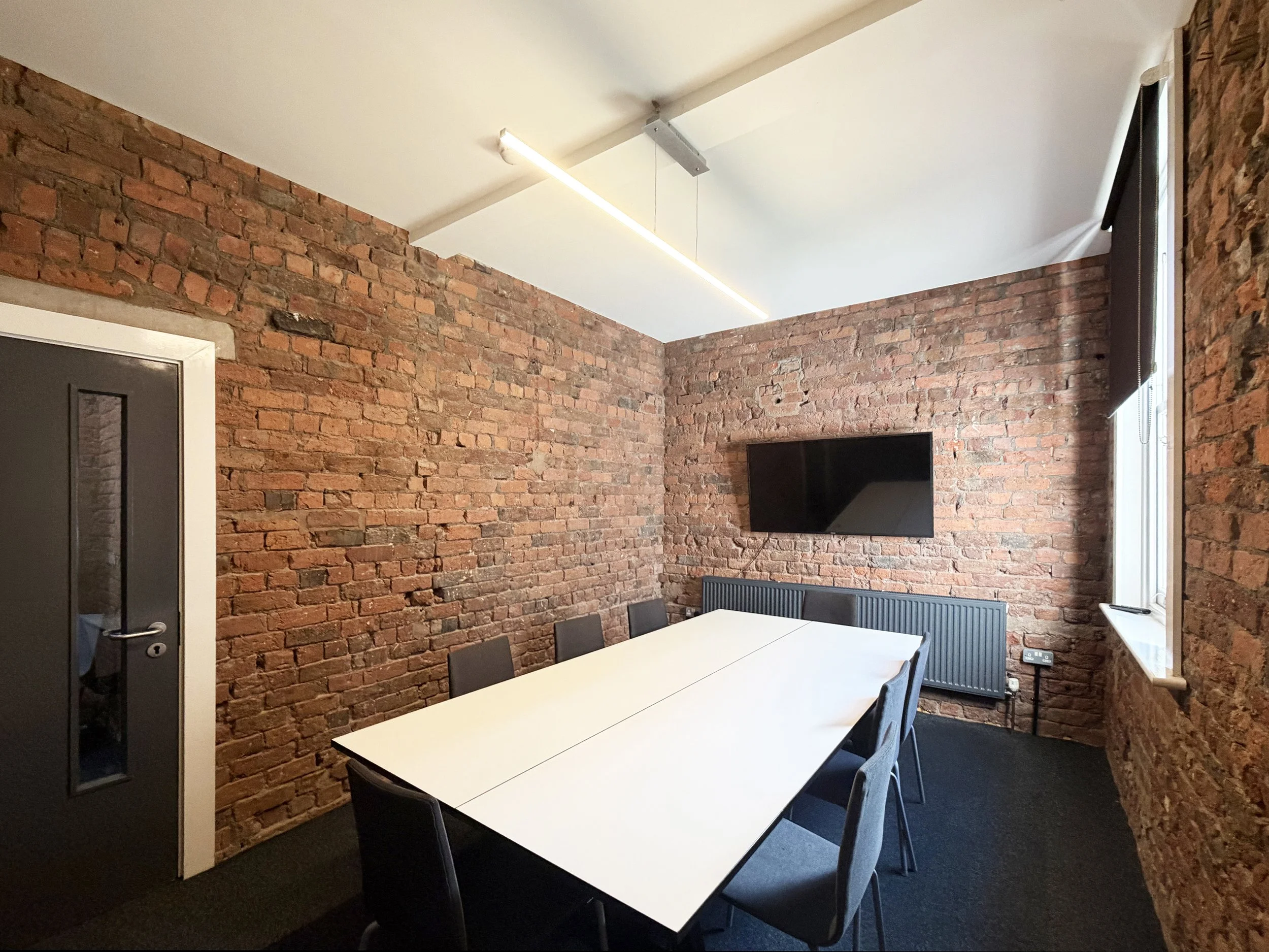 A modern conference room with exposed red brick walls, a white rectangular table, black chairs, a wall-mounted TV, a radiator, and a window with closed blinds, illuminated by a long ceiling light.