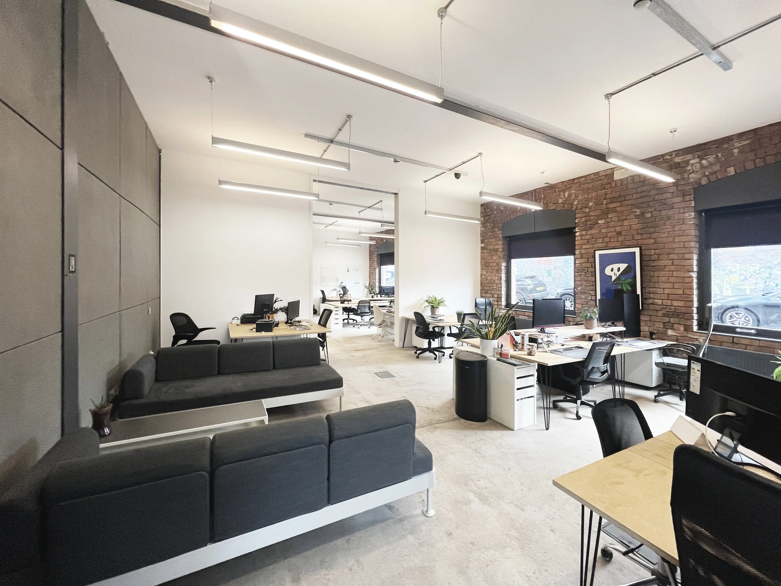 Coworking studio, Baltic Triangle  Open-plan office with multiple desks and chairs, exposed brick walls, large windows, plants, and modern hanging light fixtures.