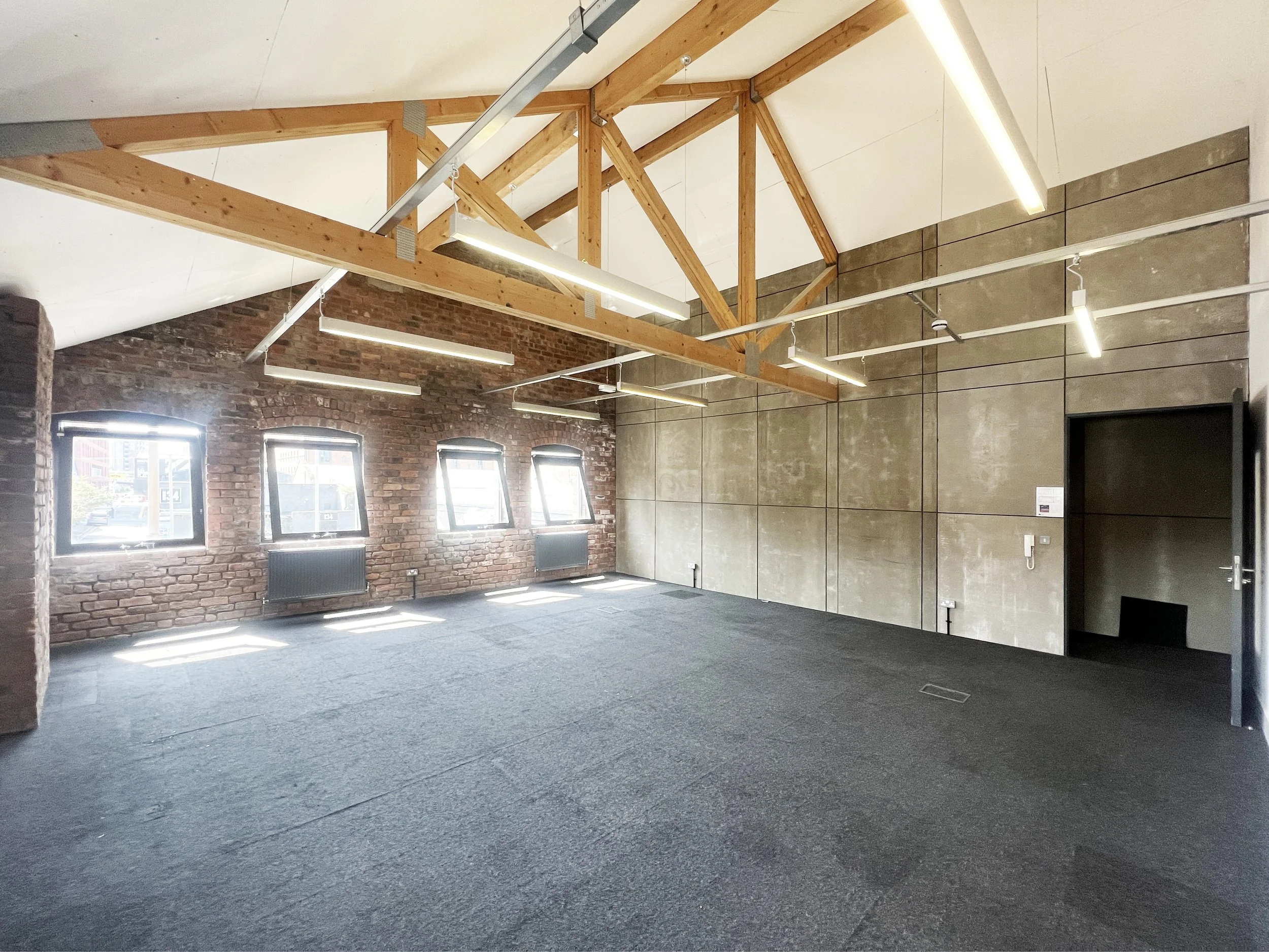 Studio Offices, Baltic Triangle Interior of an empty modern classroom or office space with exposed wooden beams, brick and concrete walls, large windows, and ceiling lights.