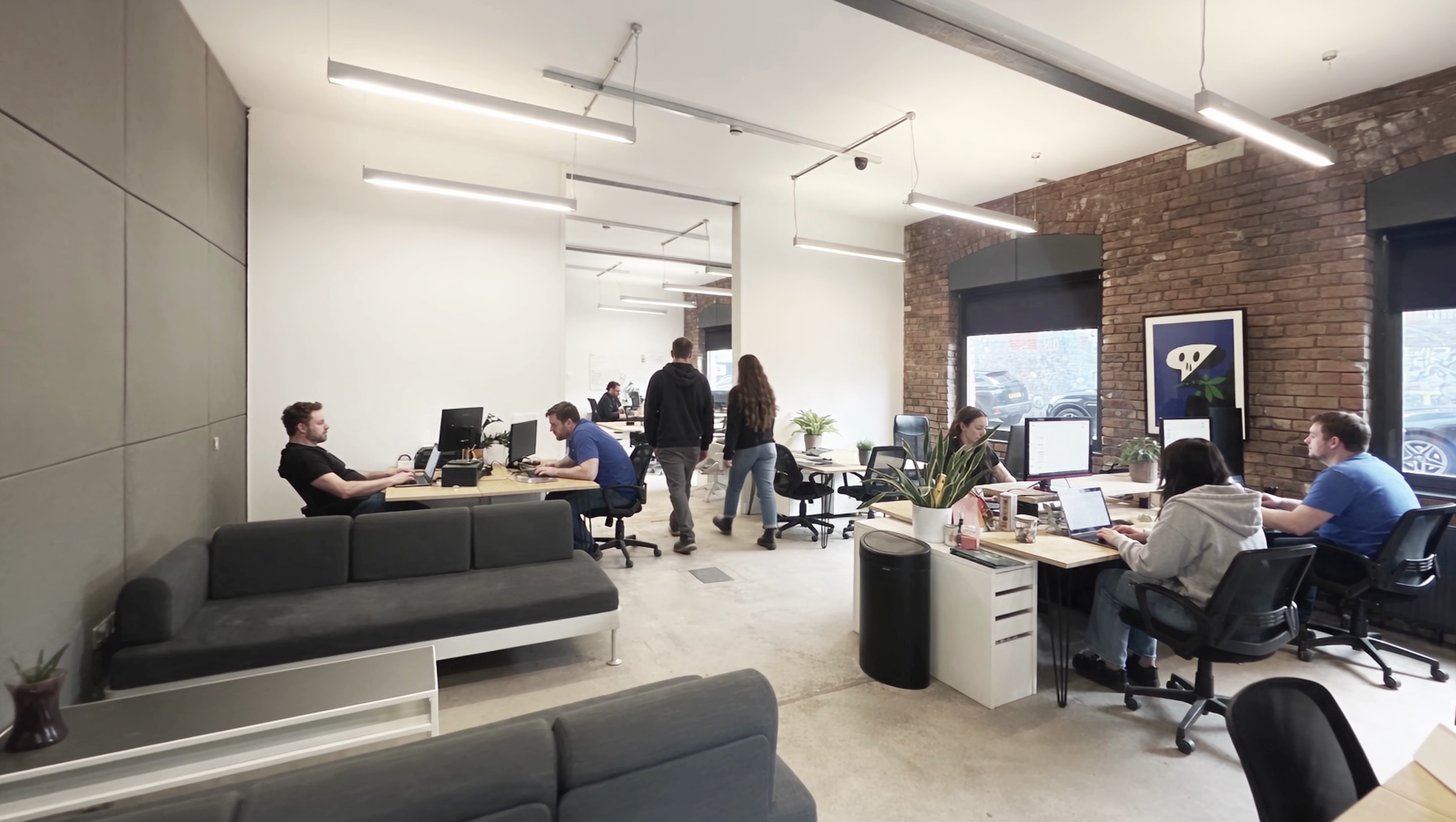 Coworking studio, Baltic Triangle  Open-plan modern office with brick and white walls, large windows, and several people working at desks with computers. Some employees are walking, and there are various plants and artwork in the space.