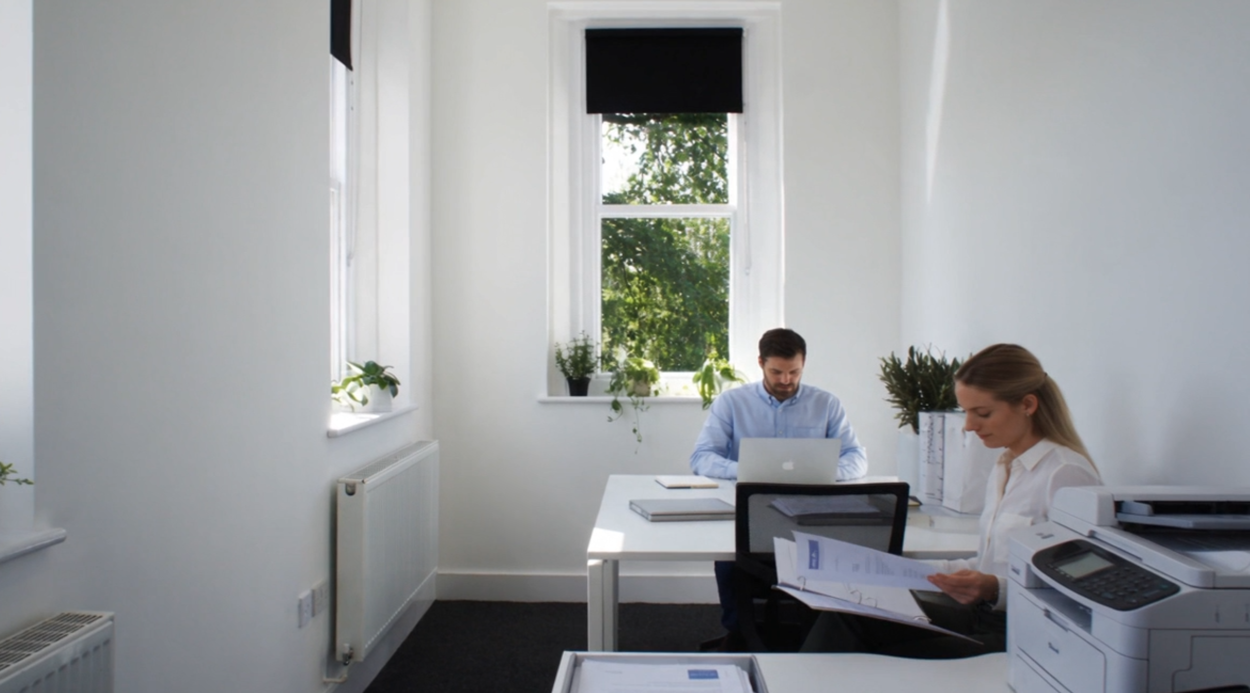 Two people in a bright, white office room working at a desk with computers, documents, and office supplies, with large windows and greenery outside.
