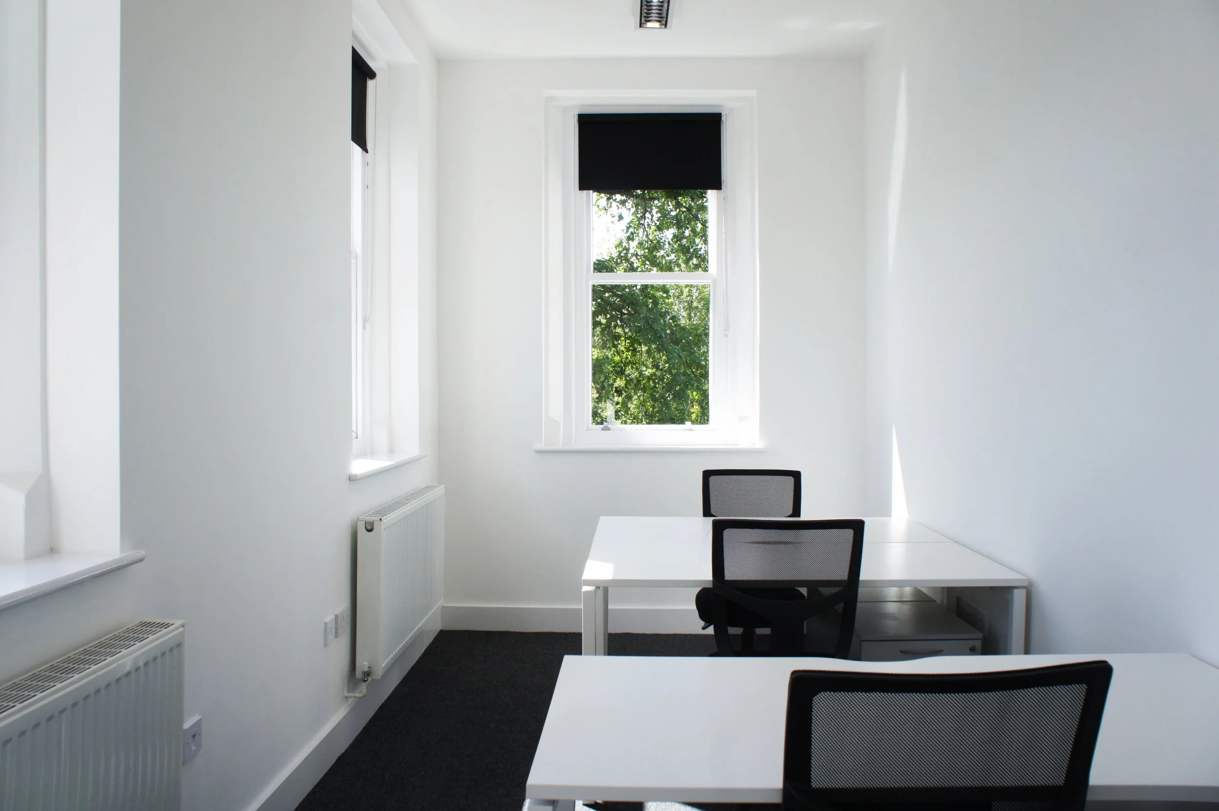 White office room with white tables and black mesh office chairs, a window with black blinds showing green foliage outside, white walls, a radiator, and a ceiling vent.