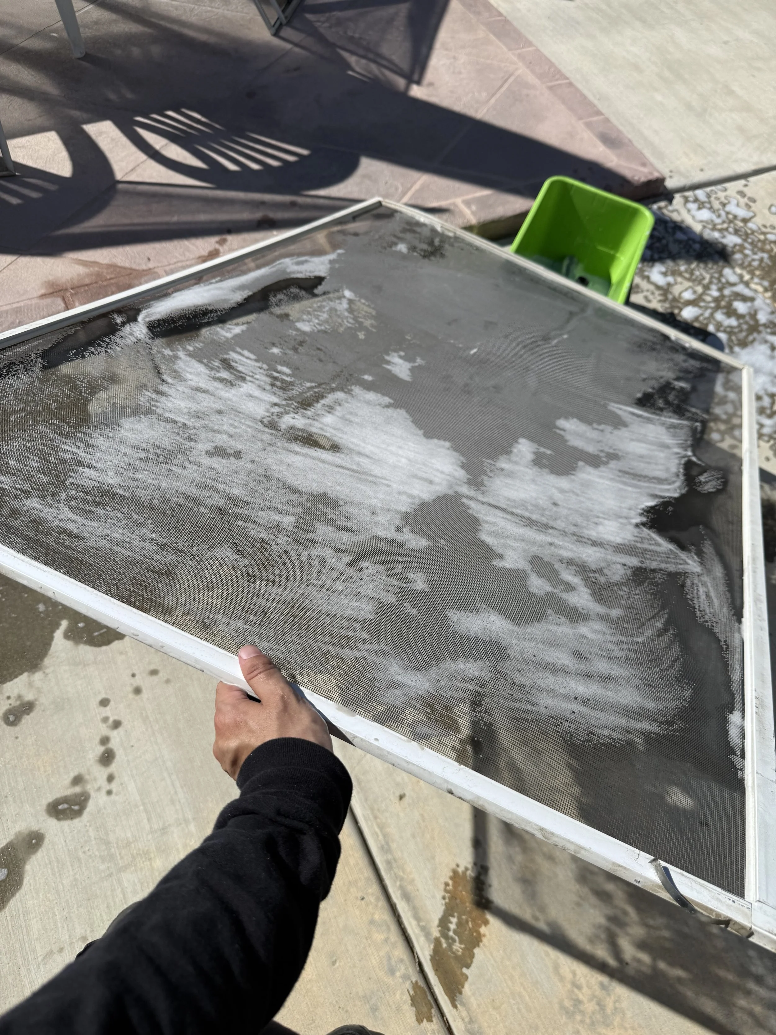 Deep scrubbing and washing a dusty mesh window screen on a concrete patio.