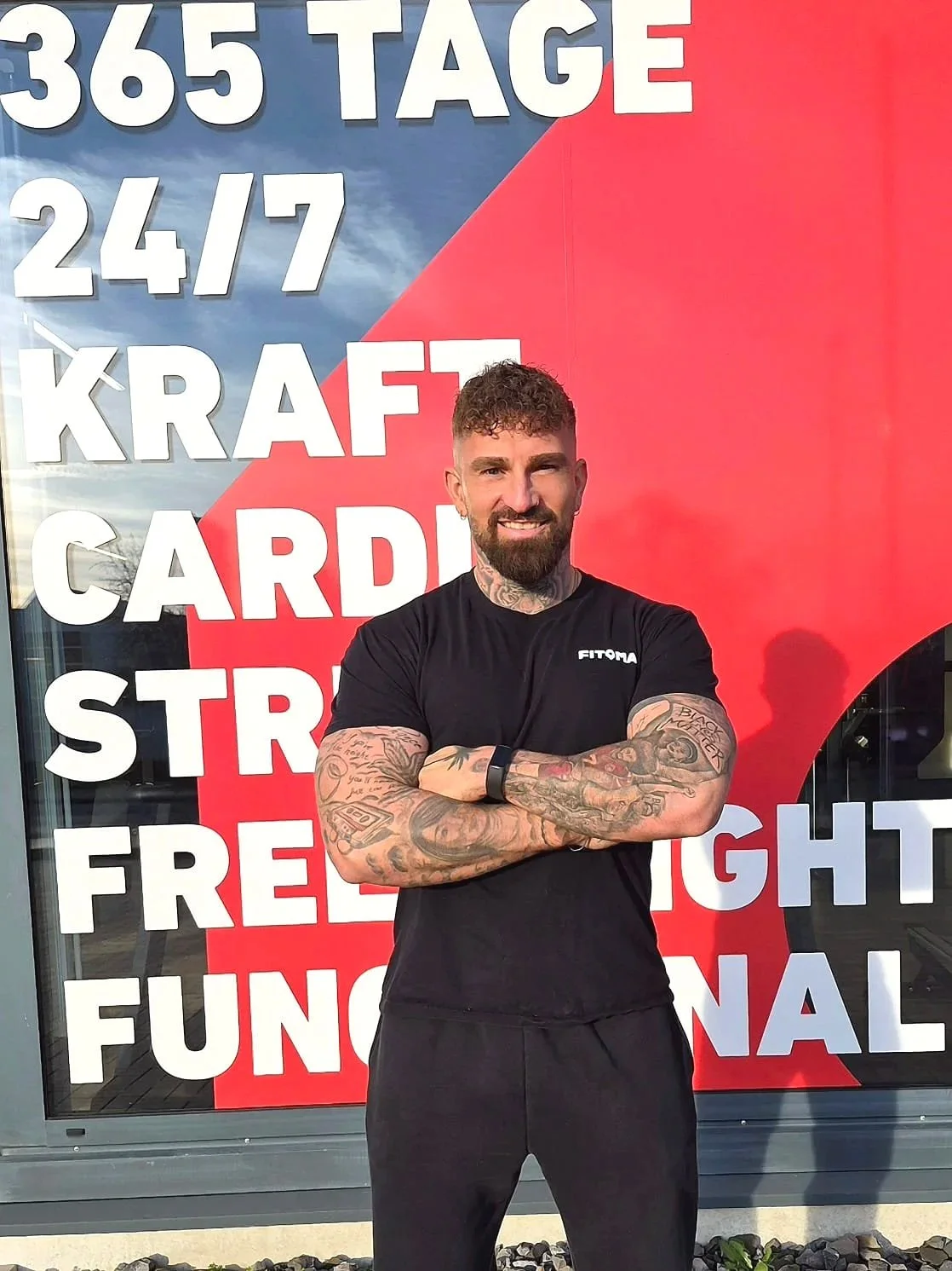 Ein muskulöser Mann mit tattoos steht vor einem Plakat mit weißen Texten auf rotem und blauen Hintergrund, der über 365 Tage, 24/7 Kraft, Cardio, Stre, Frei, Light, Funktionalität spricht.