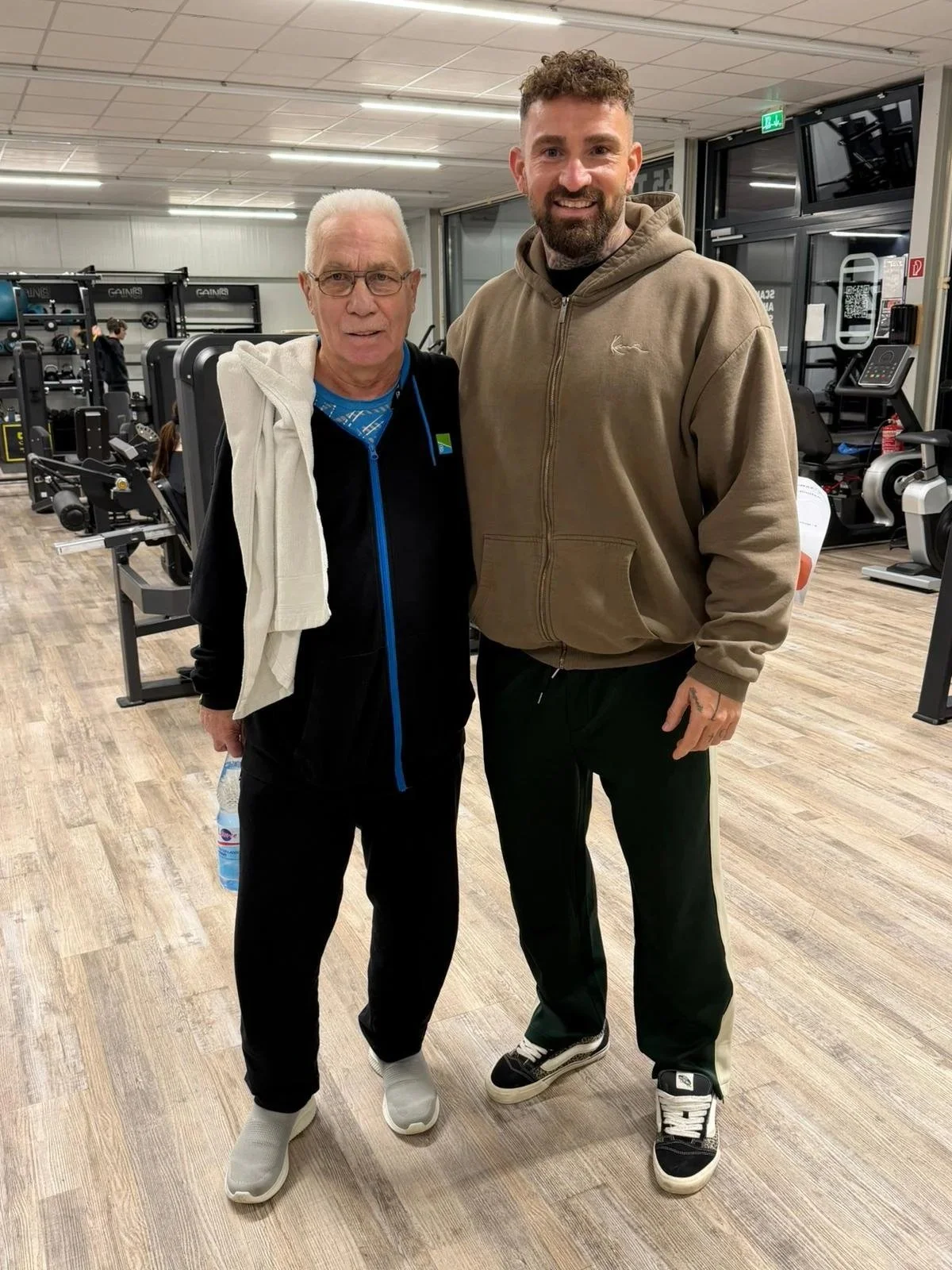 Zwei Männer stehen zusammen in einem Fitnessstudio und lächeln in die Kamera.