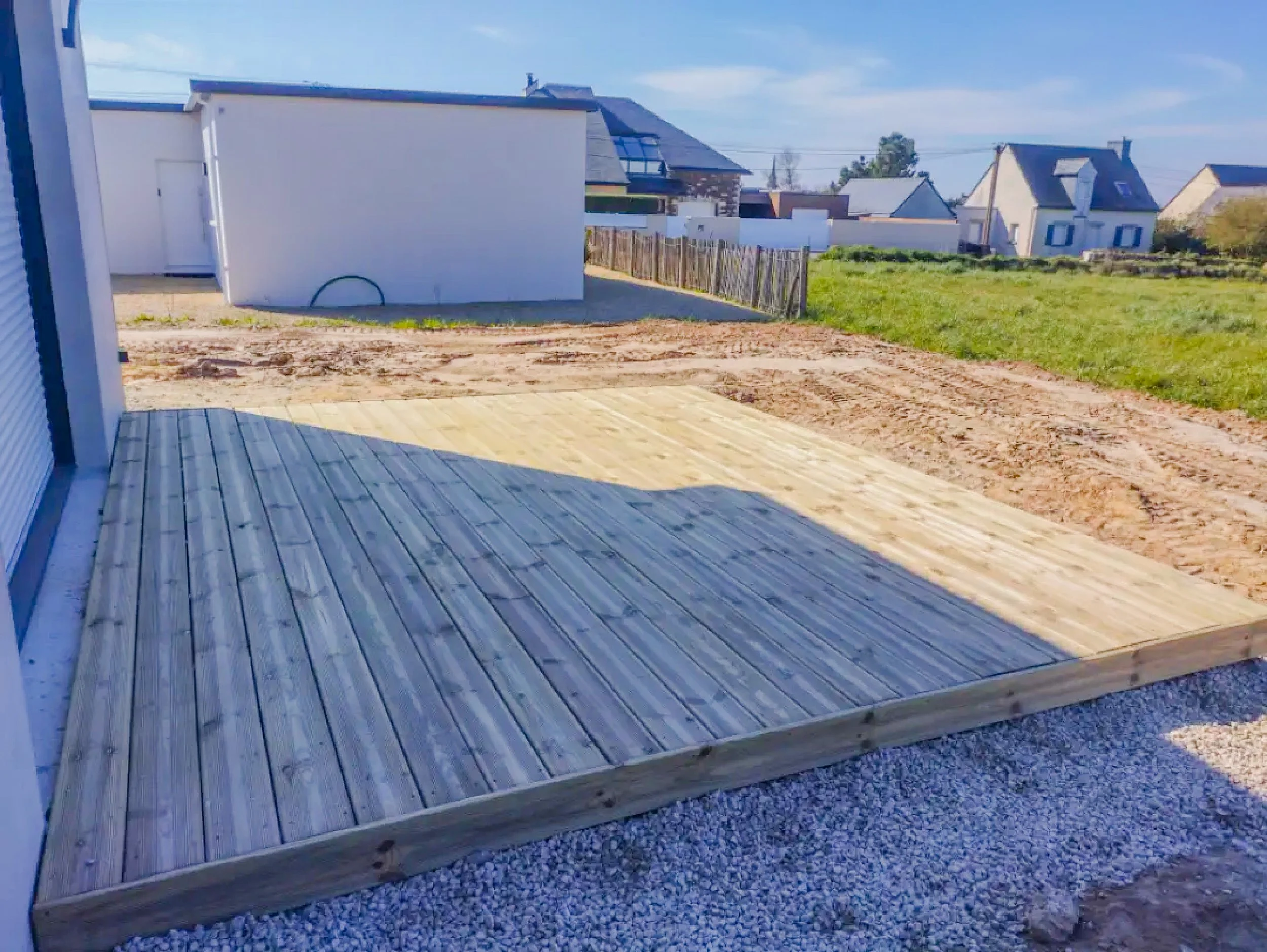 Un patio en cours de construction avec une partie en bois et une partie en terre, dans un environnement résidentiel avec plusieurs maisons en arrière-plan.