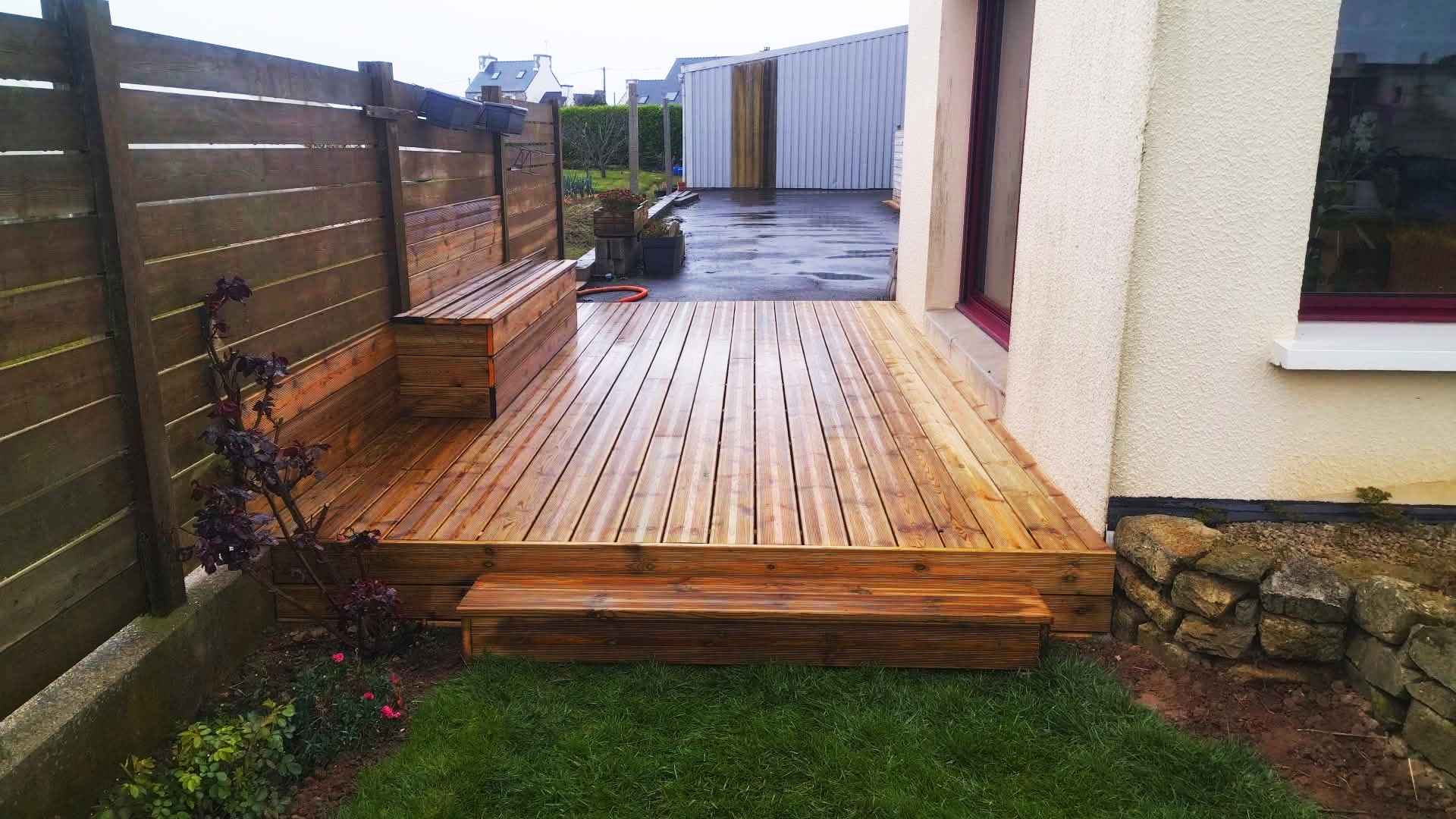 Une terrasse en bois neuve adjacente à une maison, avec un banc en bois, une petite étape, et un jardin avec des plantes et une clôture en bois. Il y a également un tuyau d'arrosage et un mur de pierres.