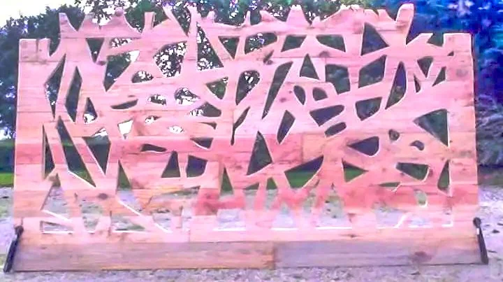 Une installation artistique en bois composée de formes géométriques découpées, placée devant un environnement naturel avec des arbres en arrière-plan.