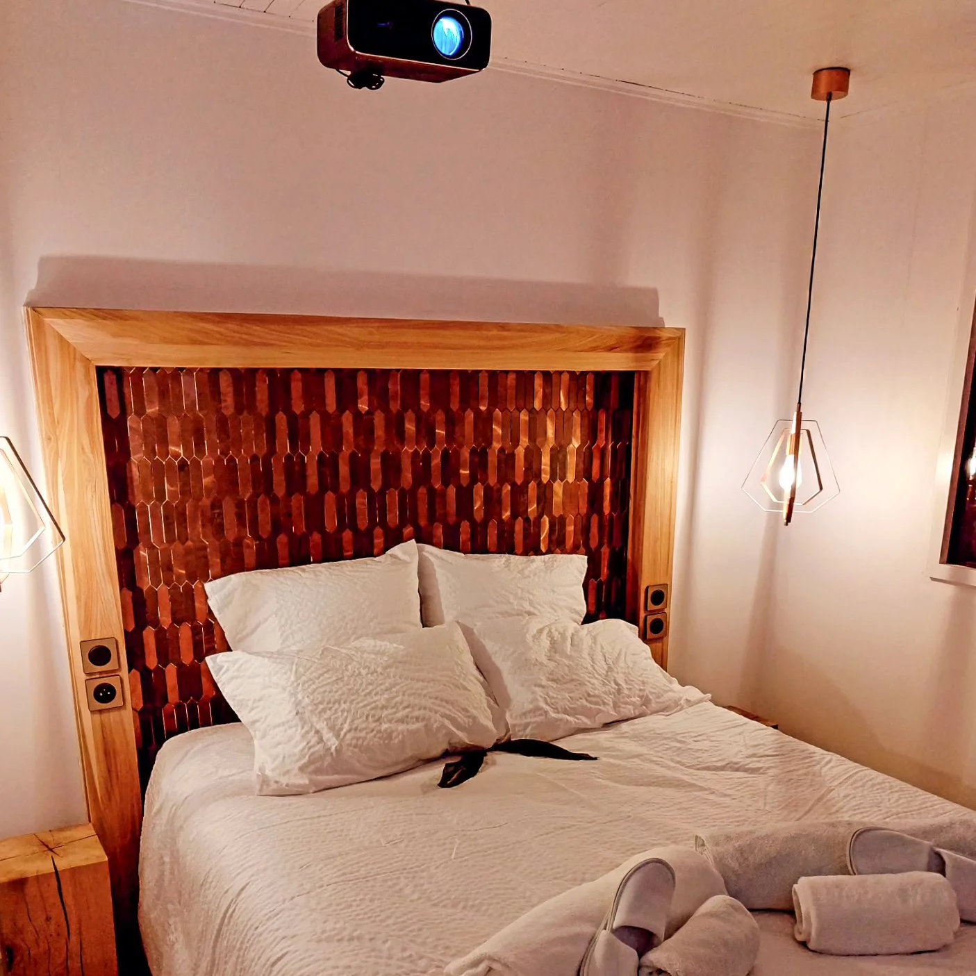 Lit avec tête de lit en bois avec motif géométrique, deux lampes suspendues à gauche et à droite, projecteur au plafond, rideau à droite, et serviettes pliées sur le lit.