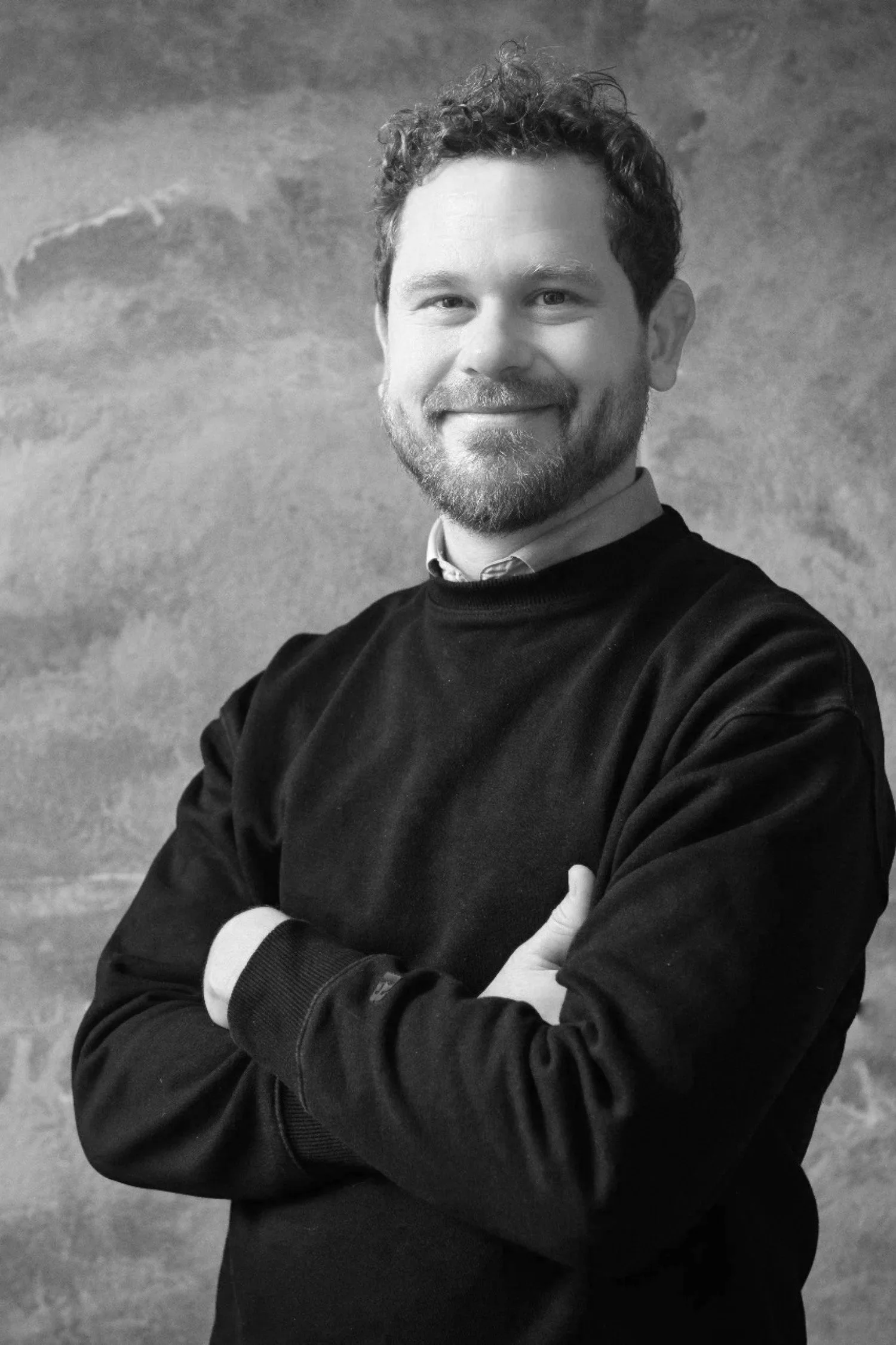 A smiling man with curly hair and a beard, wearing a dark sweater over a collared shirt, standing with his arms crossed in front of a textured wall.