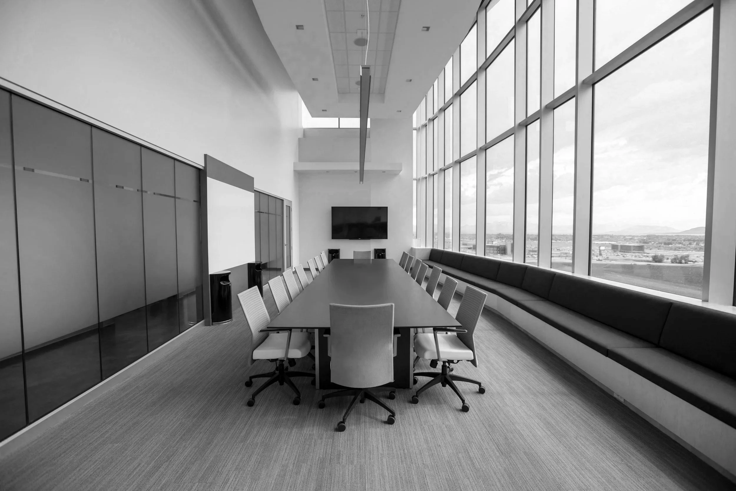 Modern conference room with large windows, a rectangular table surrounded by chairs, a wall-mounted television, and a long bench along the window.