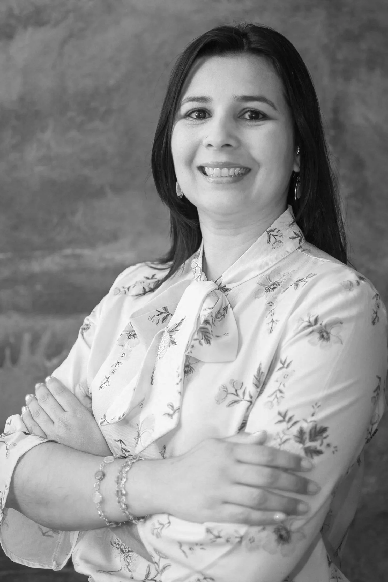 A black-and-white photograph of a woman with dark hair, smiling, wearing a floral blouse with a bow tie, earrings, and bracelets, standing against a textured background.