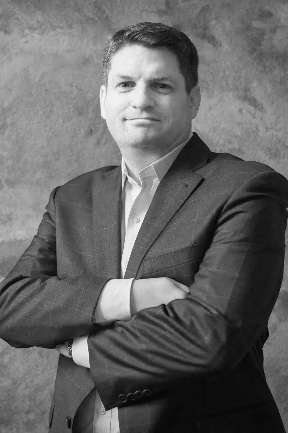 Black and white professional portrait of a man with crossed arms, wearing a suit jacket and dress shirt, standing against a textured background.
