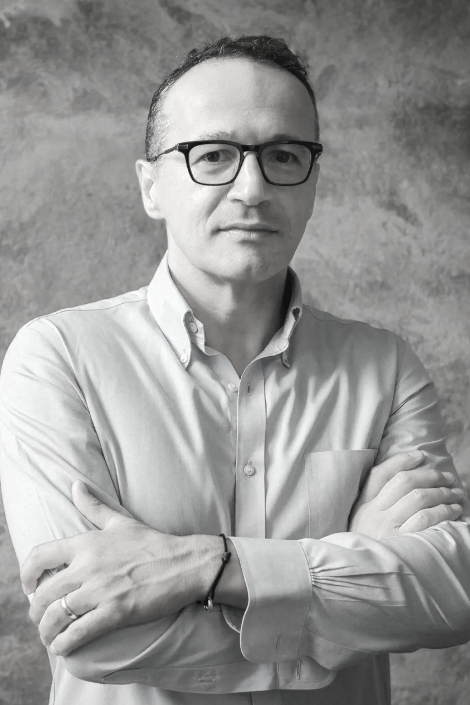Black and white portrait of a man with glasses, wearing a button-up shirt, standing with arms crossed against a textured background.