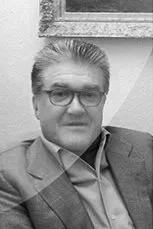 Black and white photo of a man with glasses and gray hair, wearing a suit and sitting indoors near a framed picture.