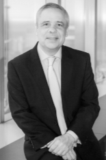 Black and white photo of a man in a suit smiling, standing with arms crossed in an indoor setting.