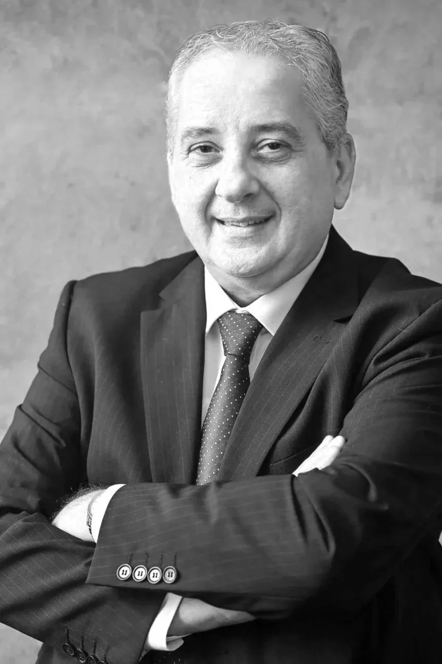 A black and white portrait of a smiling middle-aged man wearing a suit, tie, and white shirt, with arms crossed, against a textured background.