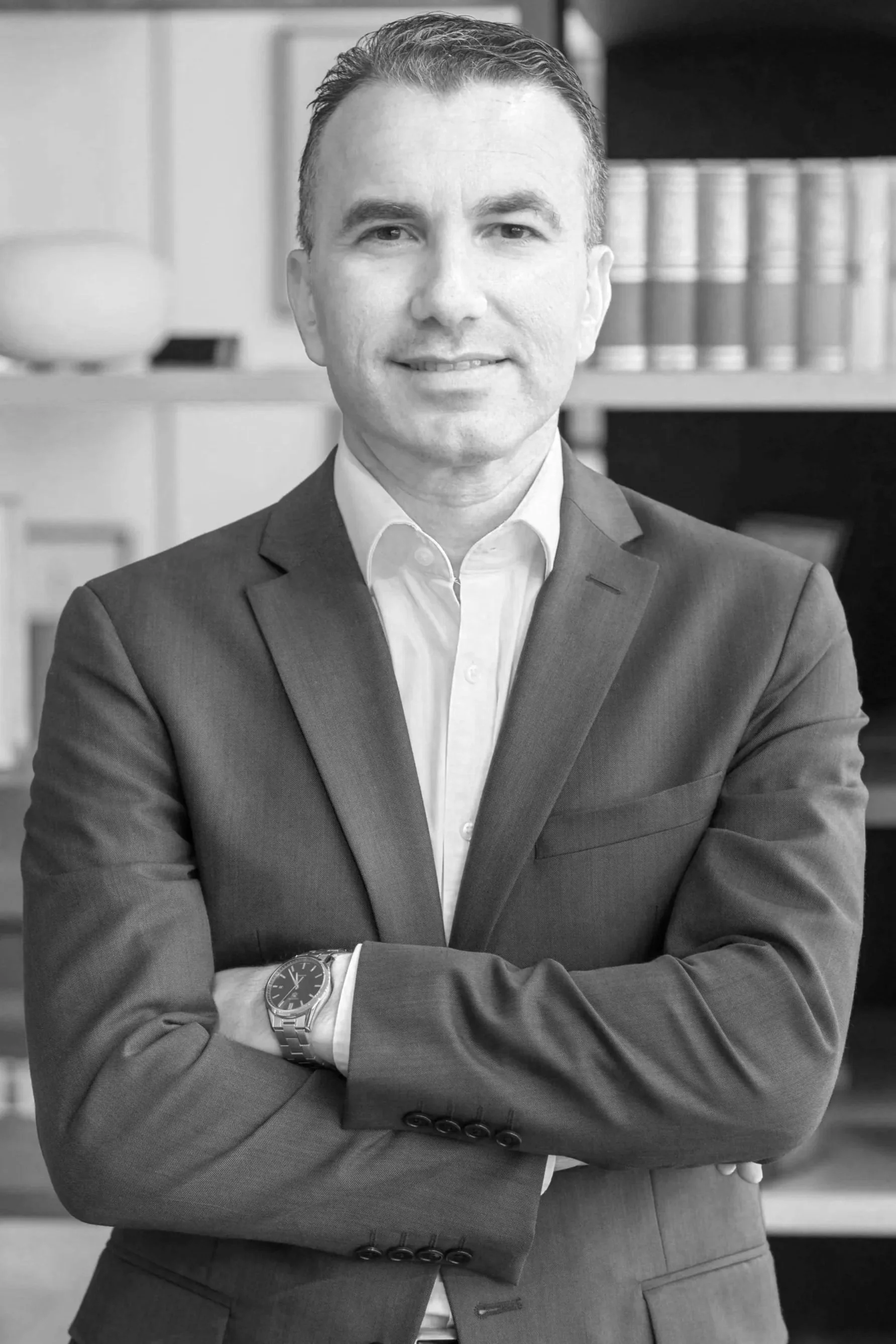 Black and white photo of a man in a suit standing with arms crossed, smiling, in an office setting with bookshelves in the background.