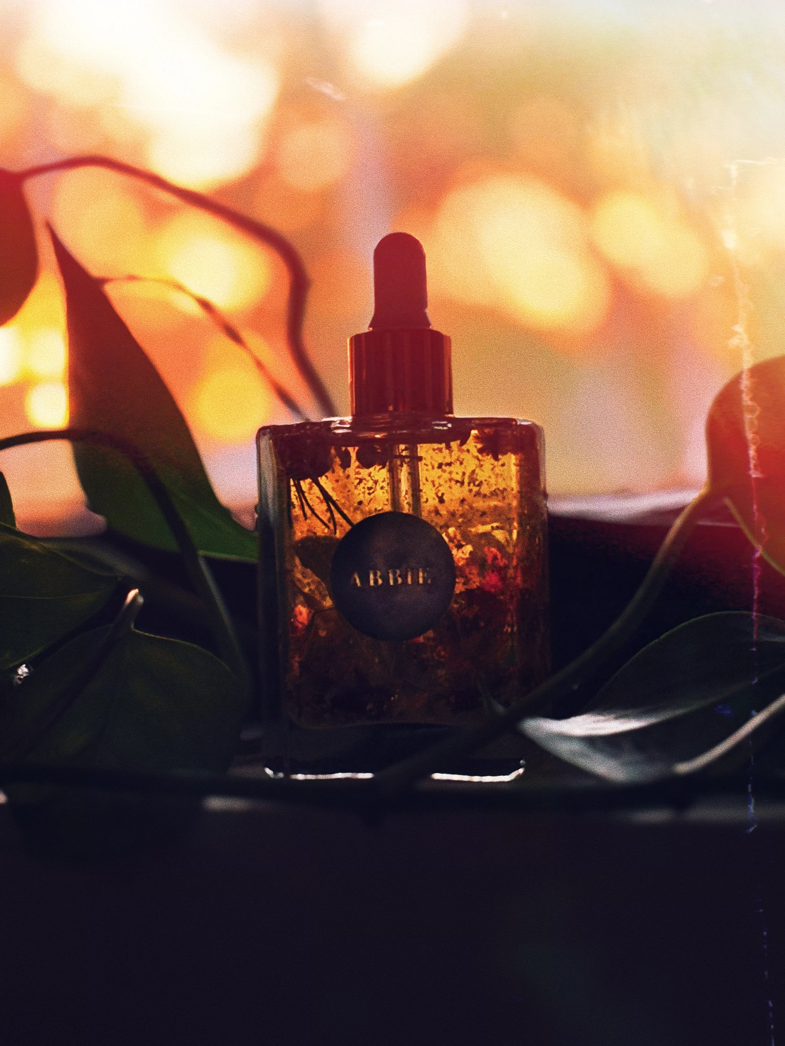 A 50mL glass dropper bottle filled with jojoba oil, herbs, botanicals, essential spell oils labelled with a personalised moon sticker that says 'ABBIE' positioned on a surface surrounded by green leaves, with a warm, blurred sunset in the background.