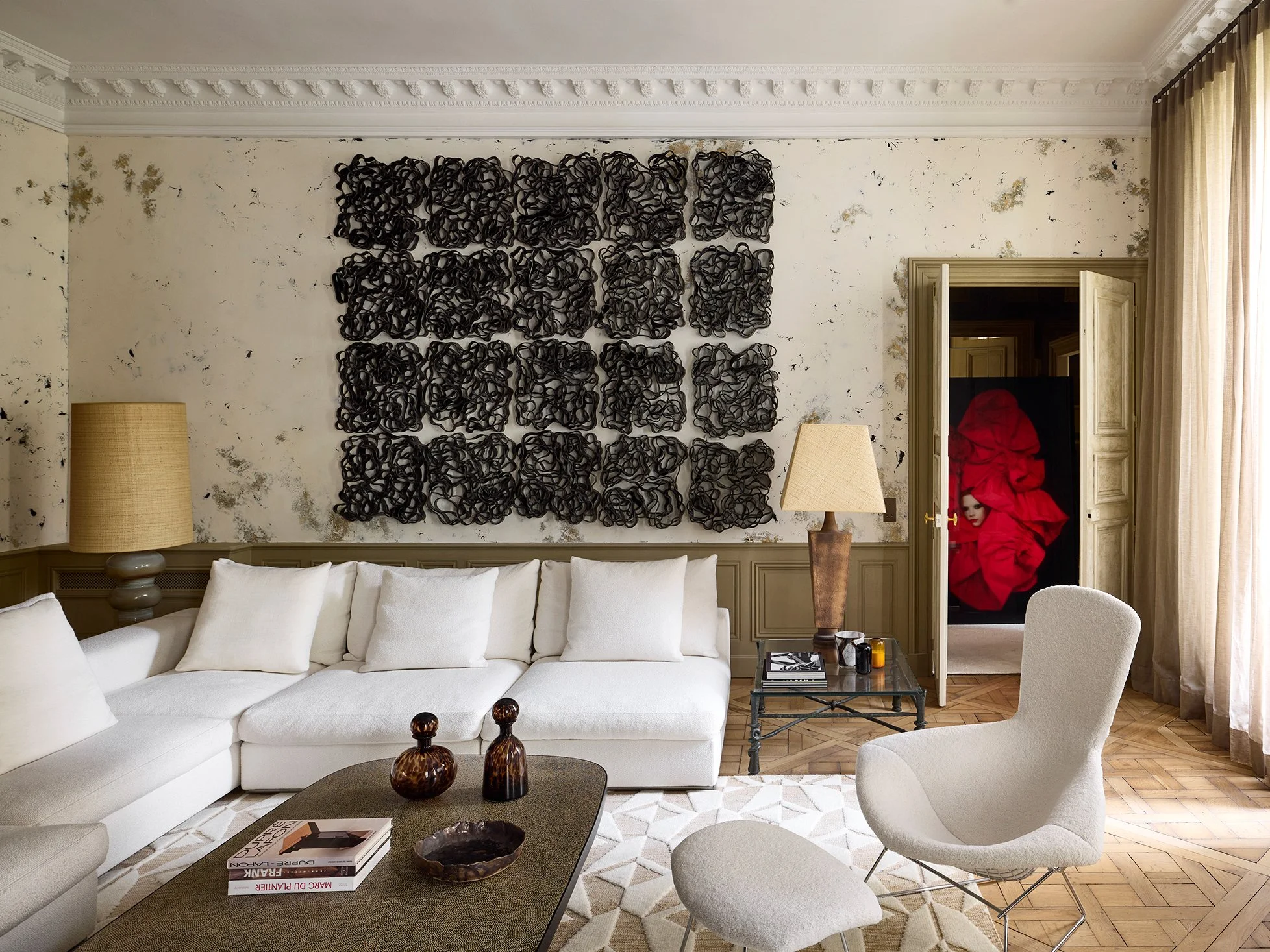 Salon moderne avec canapé blanc, table basse, chaises beige, œuvre d'art noir en relief sur le mur et porte légèrement ouverte révélant une peinture rouge.