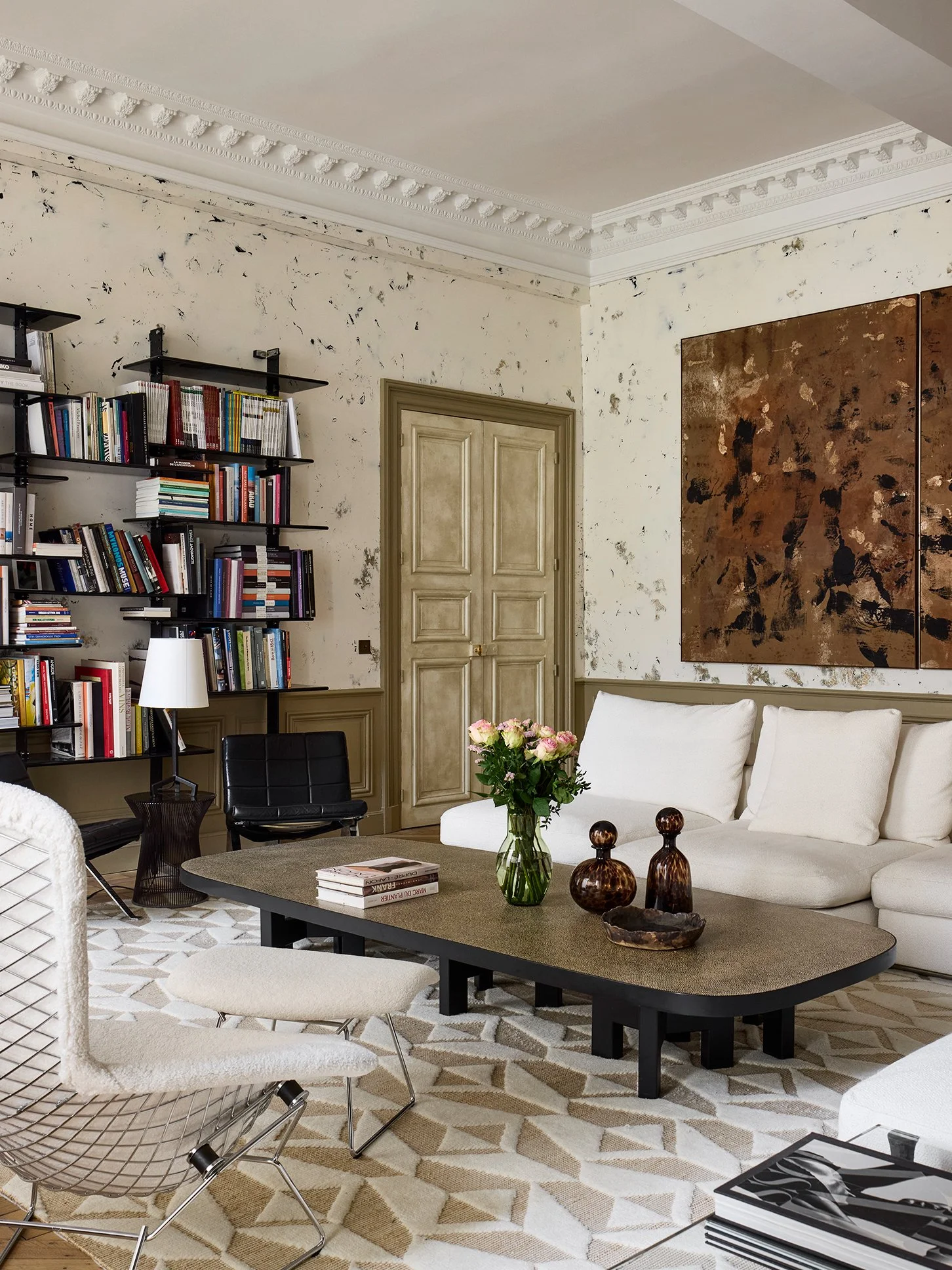 Salon élégant avec étagères remplies de livres, un sofa blanc, une table basse avec des vases et un bouquet de fleurs, un tapis géométrique beige et blanc, et une peinture abstraite murale.
