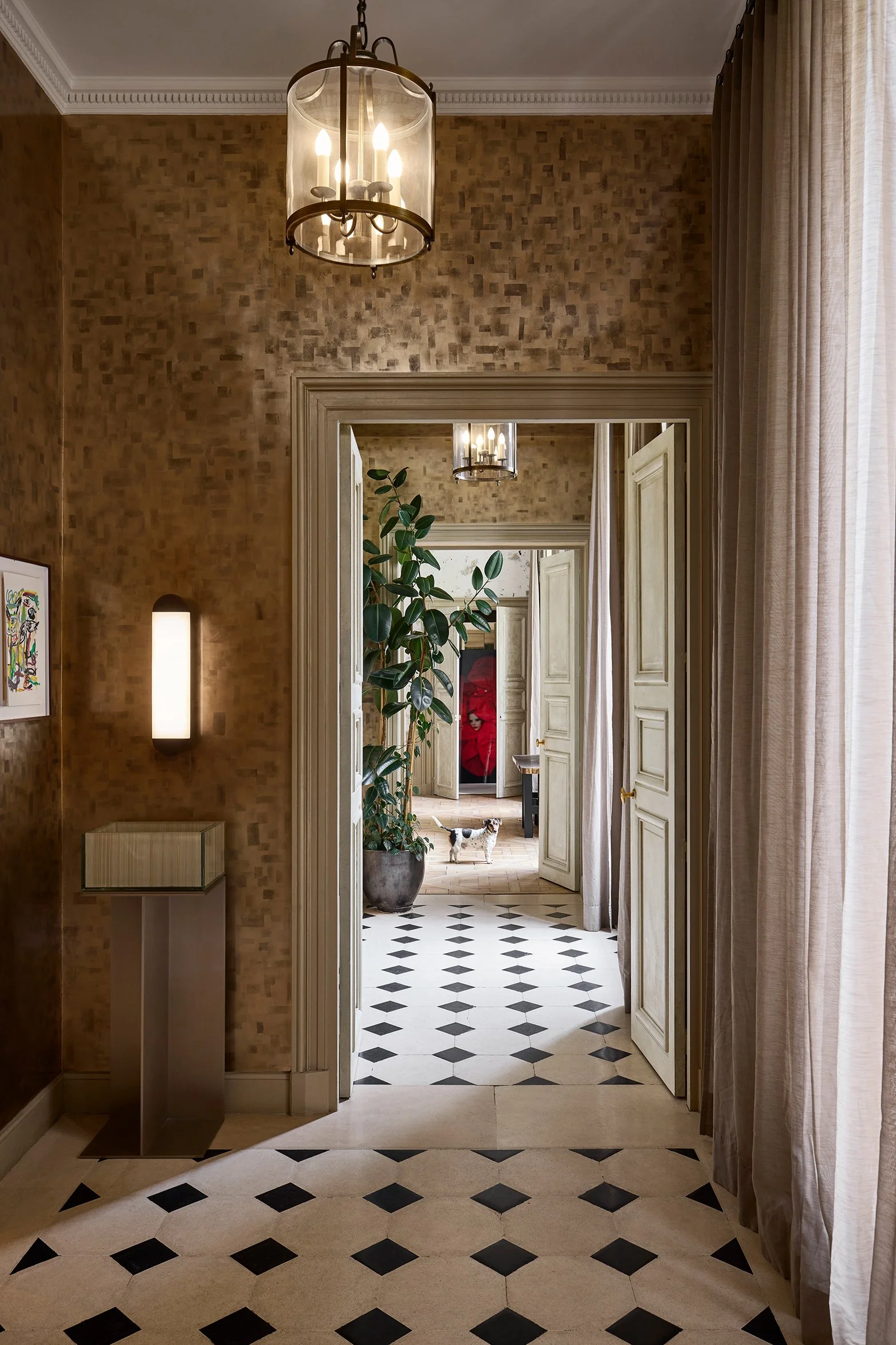 Une vue à travers plusieurs portes d'une pièce élégante avec un sol en carreaux noirs et beige, un mur beige, un grand potted plant, un chien, des rideaux clairs, un lustre au plafond