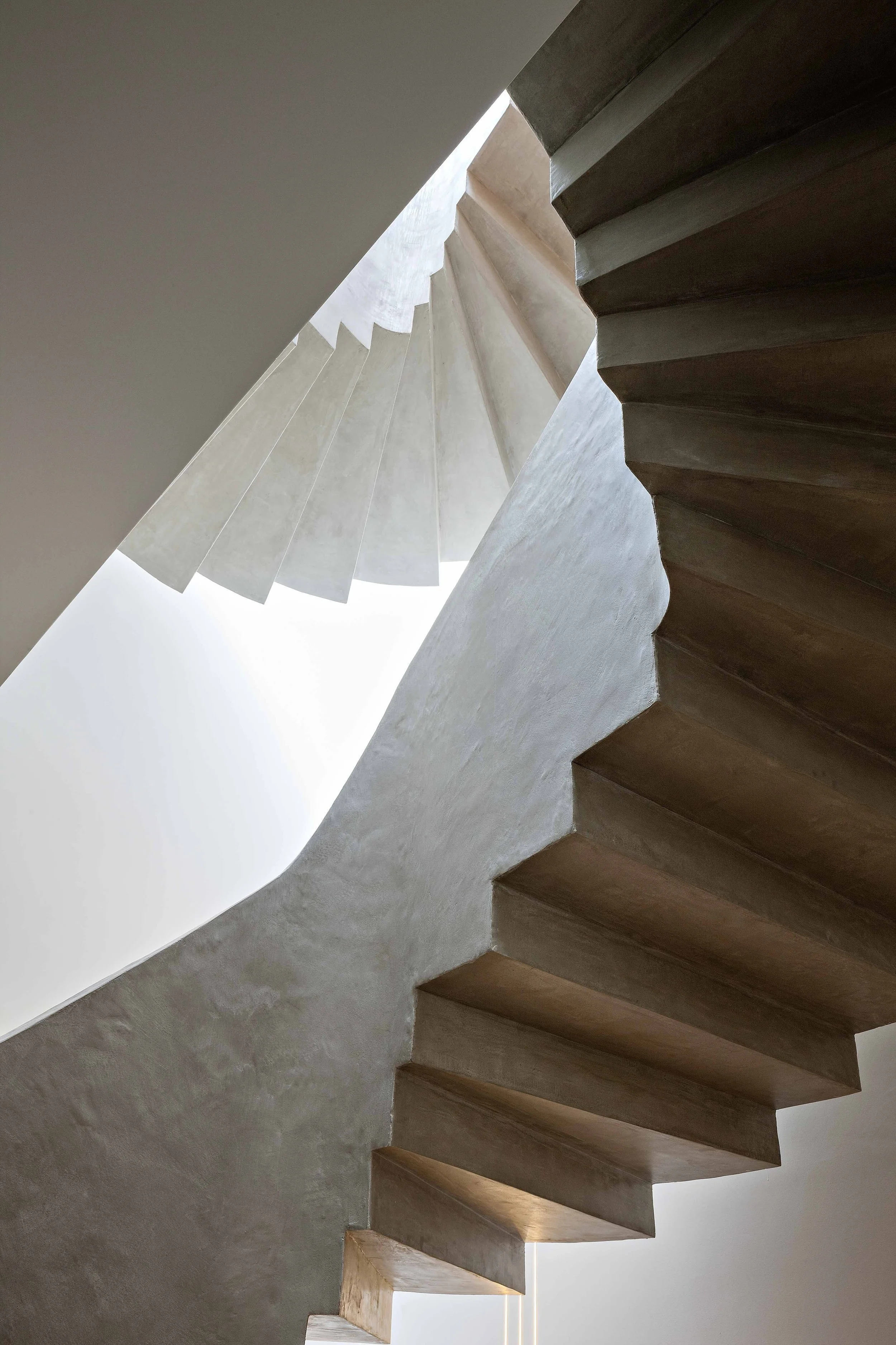 Vue en contre-plongée d'un escalier moderne en béton et bois, avec un puits de lumière au sommet.