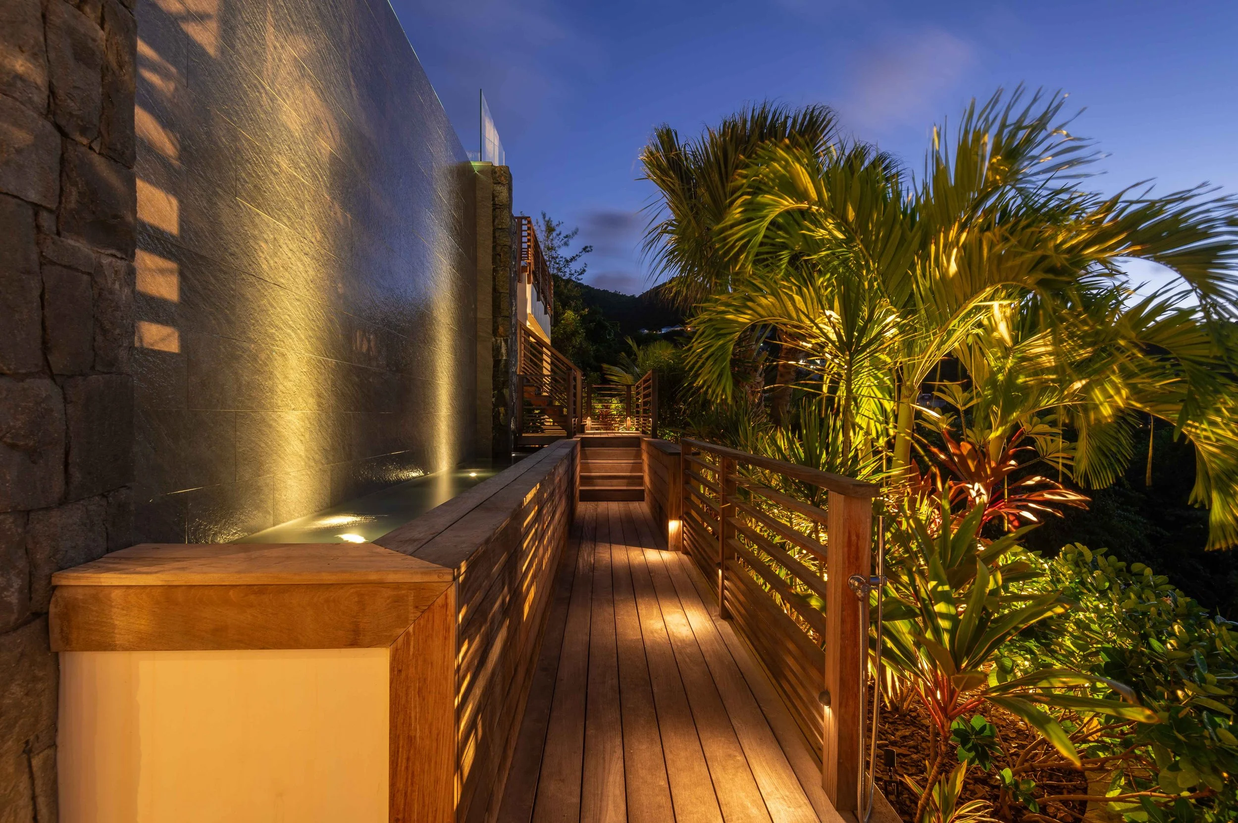 Balcon en bois avec rambarde en métal, entouré de plantes tropicales, éclairé par des lumières, au crépuscule ou nuit, vue sur la montagne en arrière-plan.