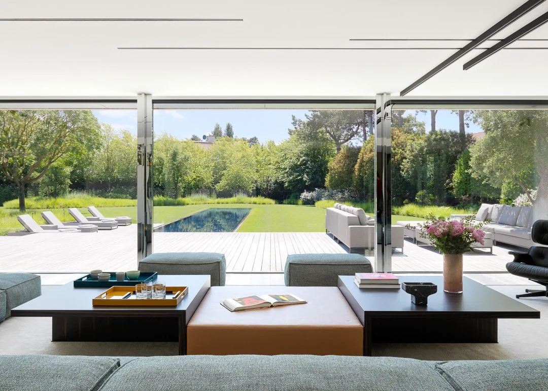 Salon moderne avec vue sur un jardin et une piscine extérieure, meubles en blanc et gris, fleurs dans un vase