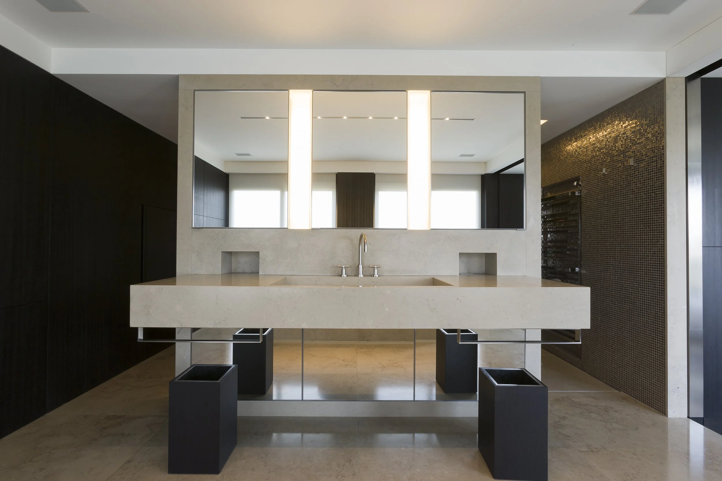 Une salle de bain moderne avec un grand miroir au-dessus du lavabo beige, un éclairage vertical intégré dans le miroir, et un mur en mosaïque dorée à droite.