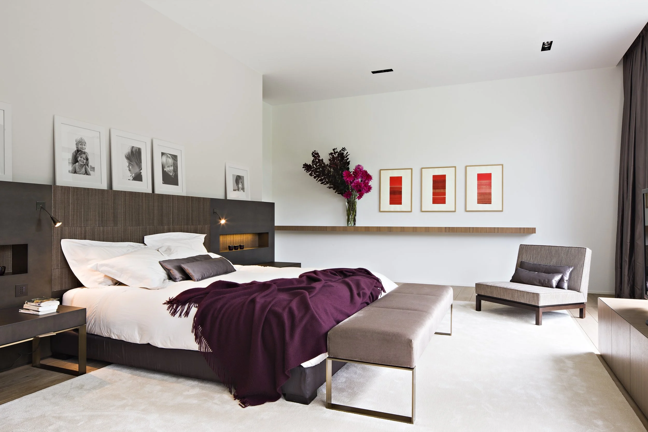 Chambre d'hôtel moderne avec lit king-size, coussins blancs et noir, couverture prune, fauteuil gris, étagère en bois avec vase de fleurs roses et rouges, trois œuvres d'art rouges encadrées, photos en noir et blanc de visages d'enfants au mur, grand