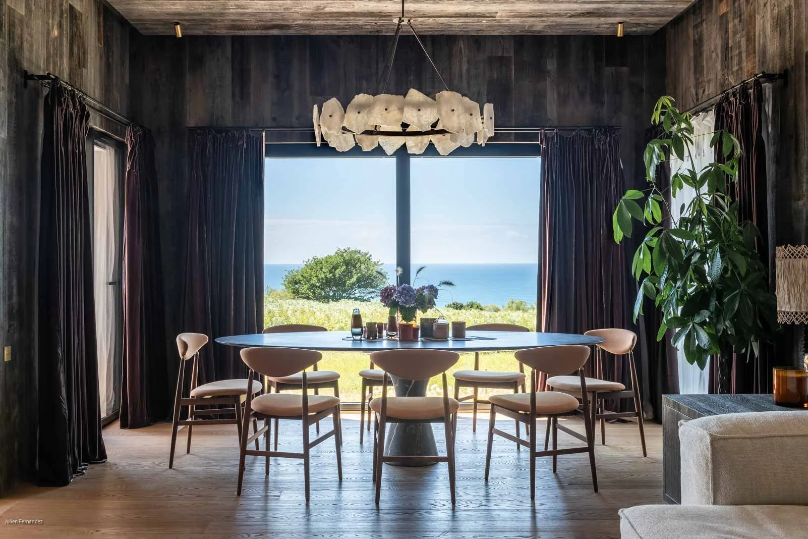 Salon avec grande fenêtre offrant une vue sur la mer, table ronde avec six fauteuils, végétation verte, lampadaire, et chandelier blanc suspendu.