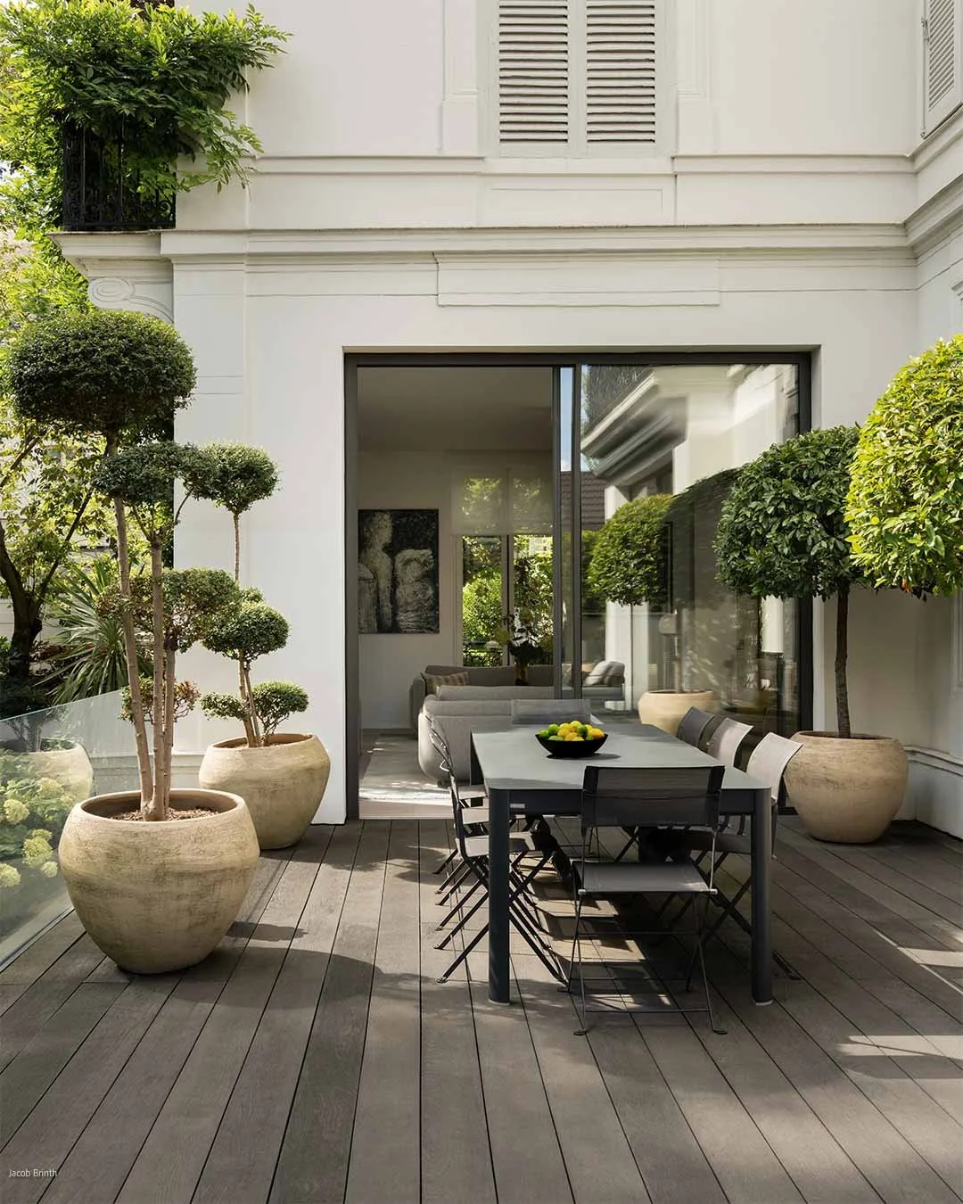 Terrasse moderne avec mobilier en bois et fauteuils noirs, entourée de grands pots avec des arbres taillés en forme, donnant sur un intérieur visible à travers une porte en verre.